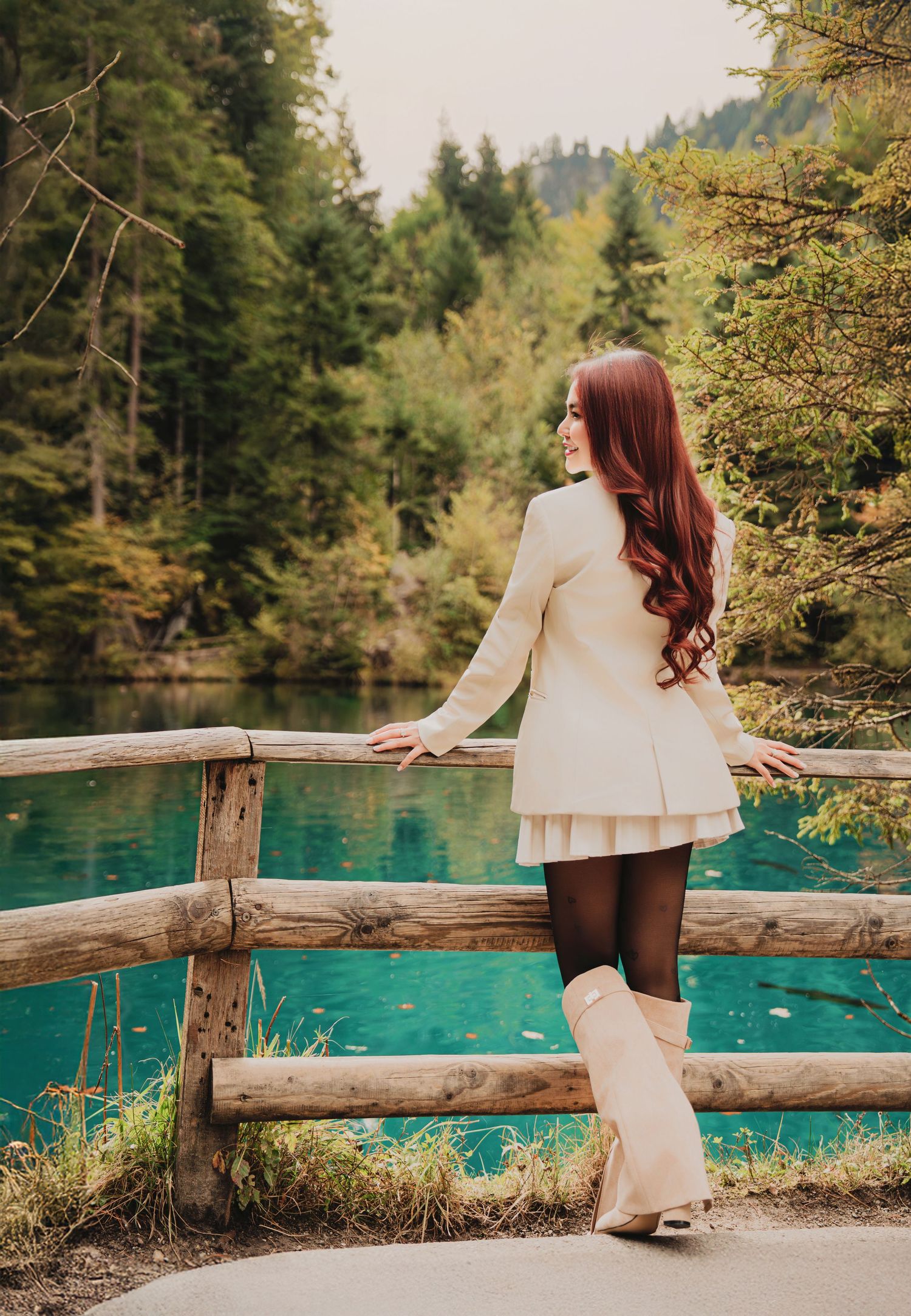 Elegant red-haired woman in white blazer and skirt posing by wooden fence at turquoise lake.