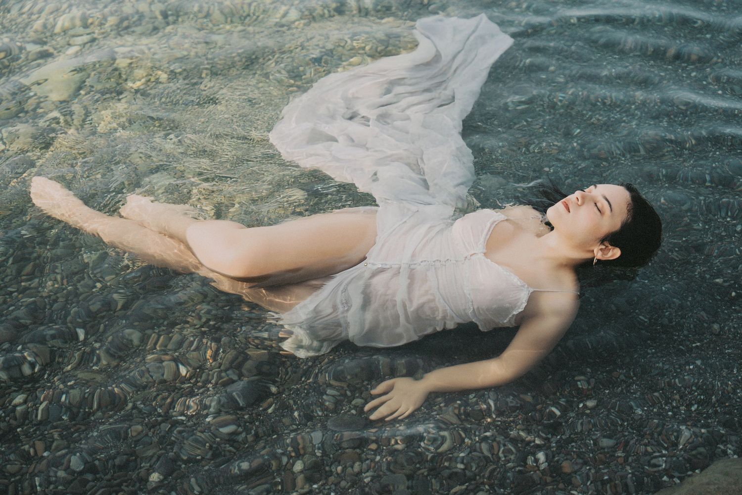 Woman in white sheer dress floating peacefully in clear shallow water.