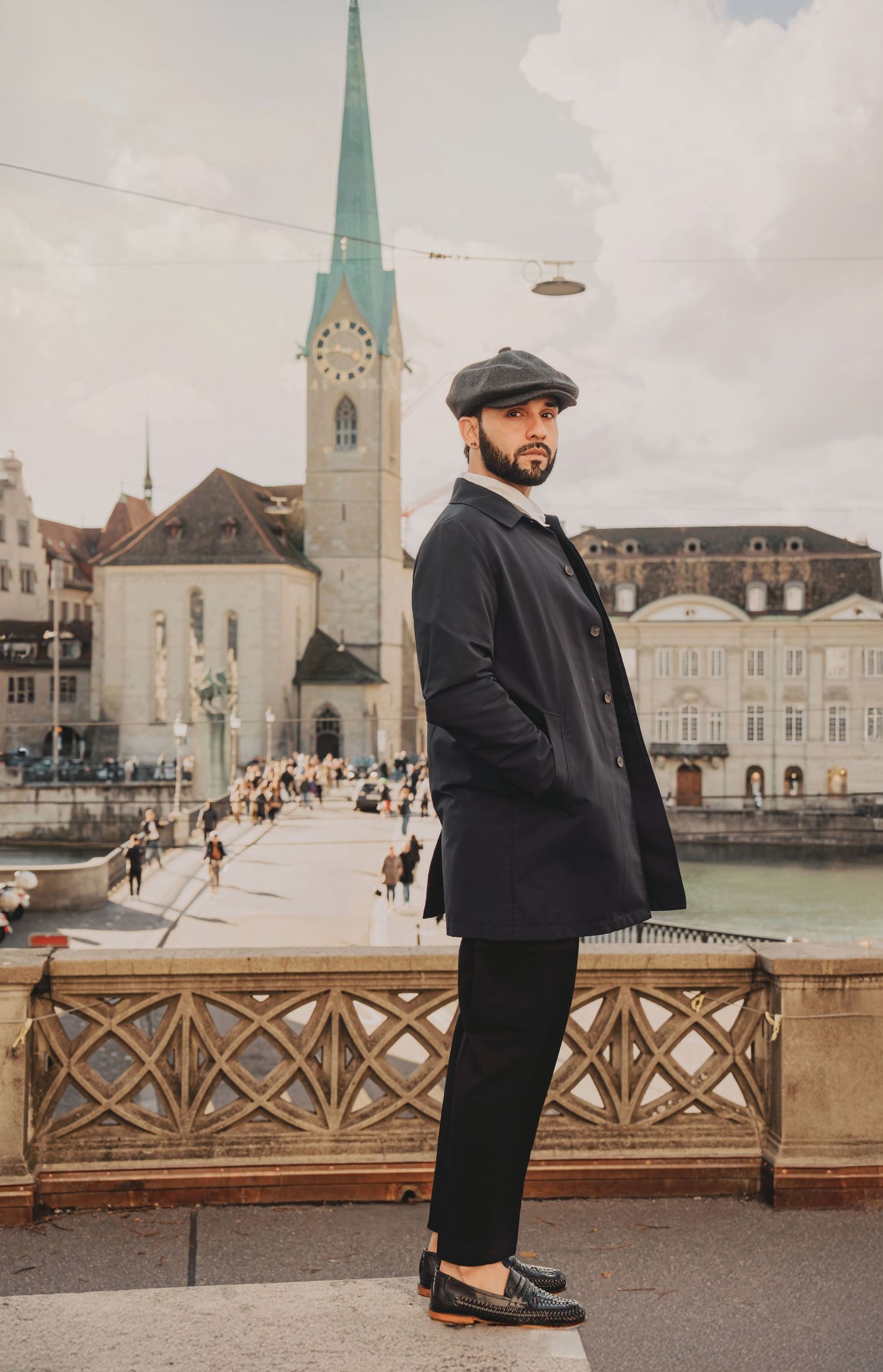 Stylish man in black coat and newsboy cap poses on Zurich riverside bridge with Fraumunster church tower.