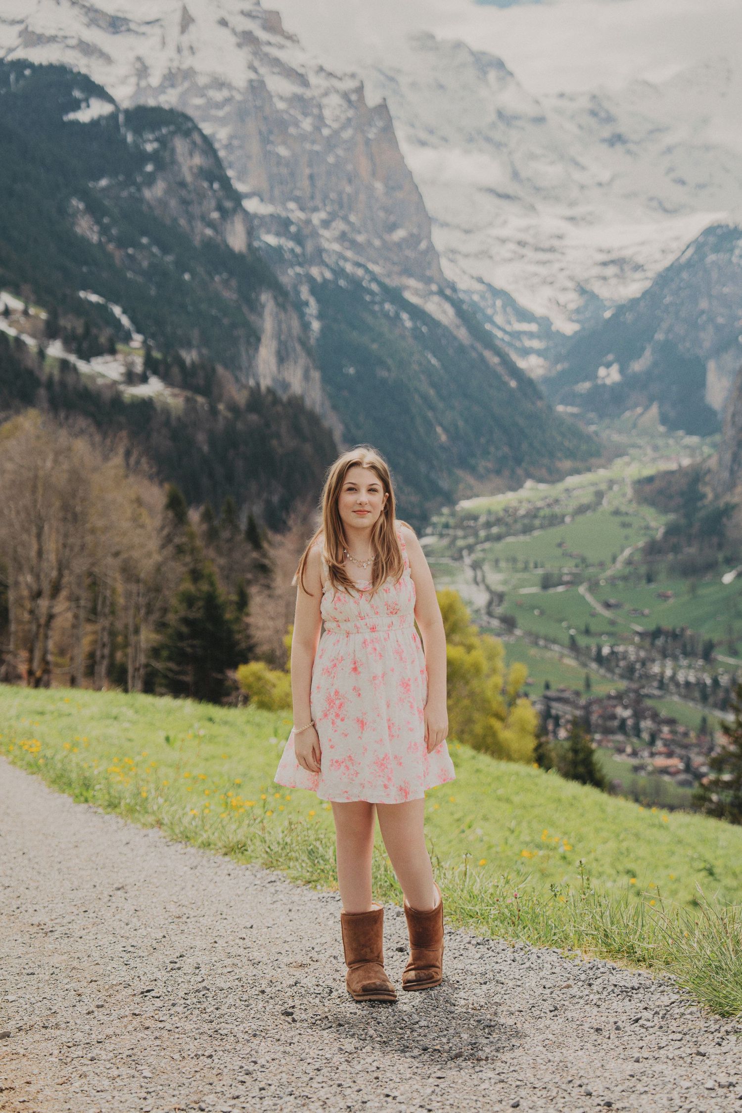 Young girl in pink floral dress and brown boots stands on mountain path in Swiss Alps valley.