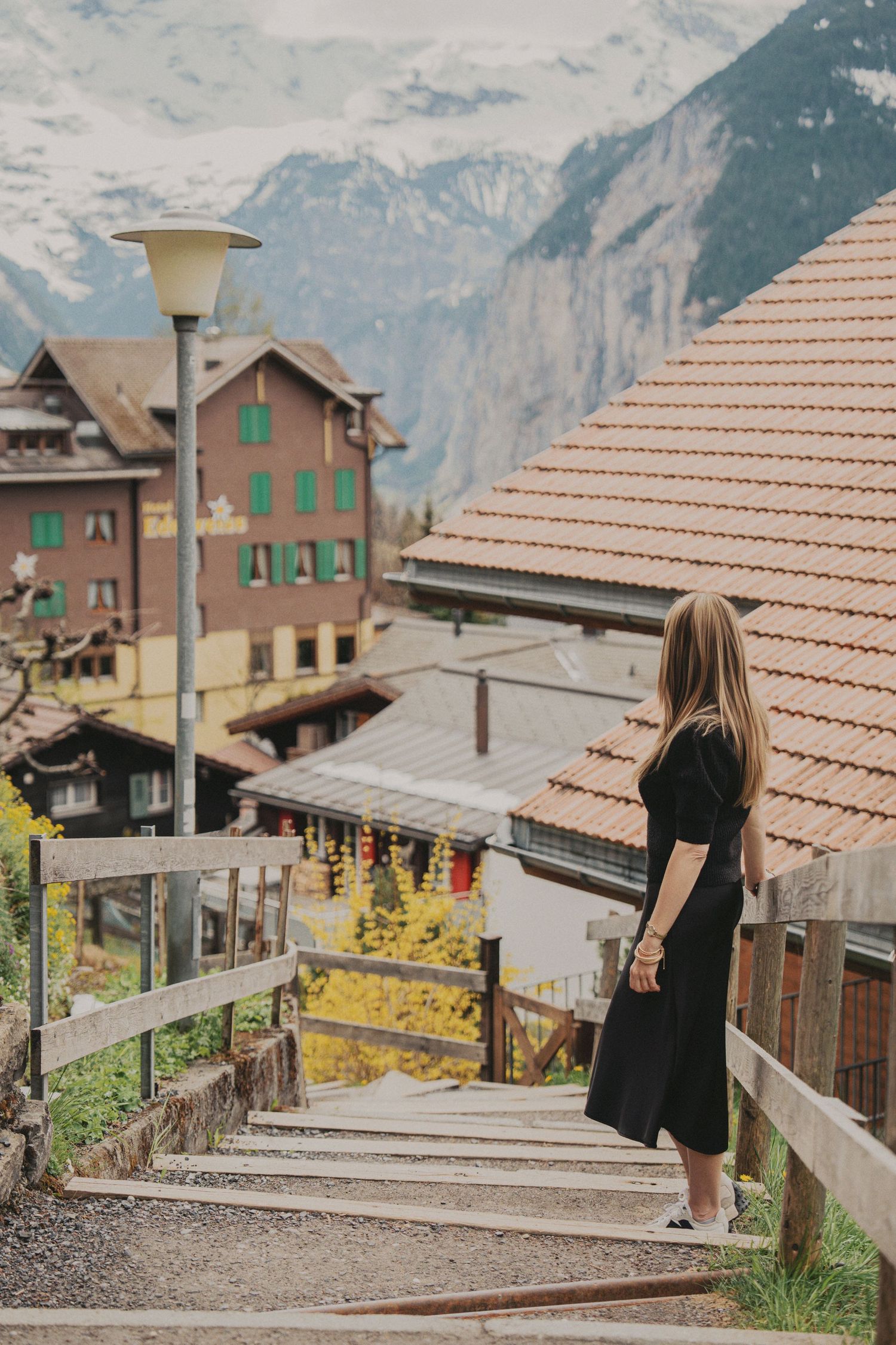 Woman in black dress standing on wooden stairs overlooking a scenic Swiss Alpine village.