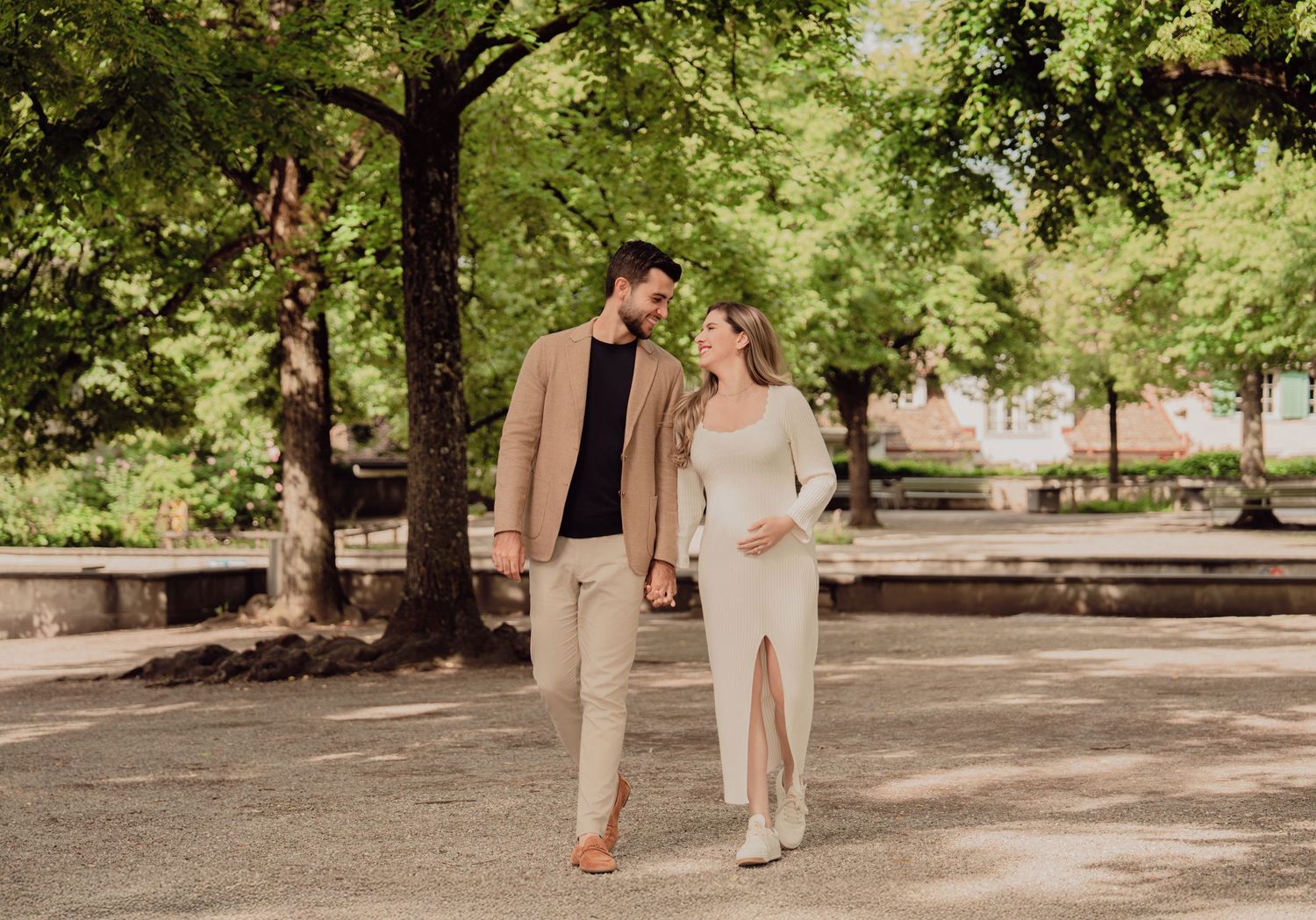 Pregnant couple strolling through a tree-lined park, woman in white maternity dress, man in tan blazer and black shirt.