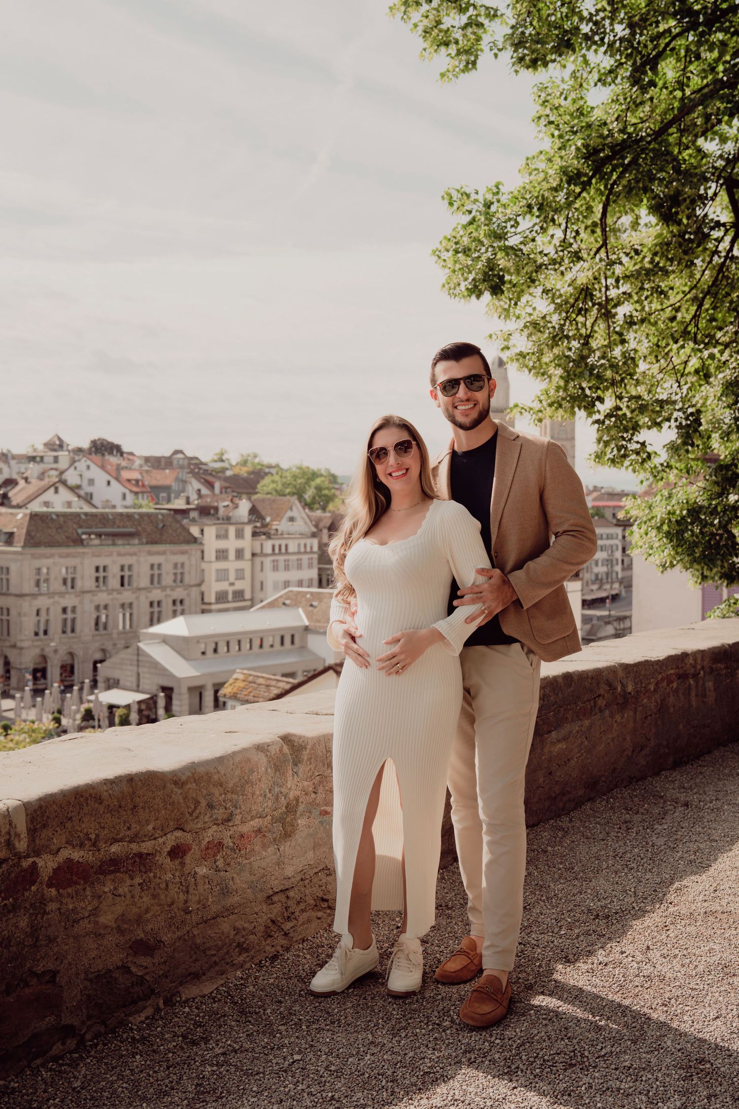 Smiling pregnant couple poses in front of European cityscape, both wearing sunglasses and neutral outfits.