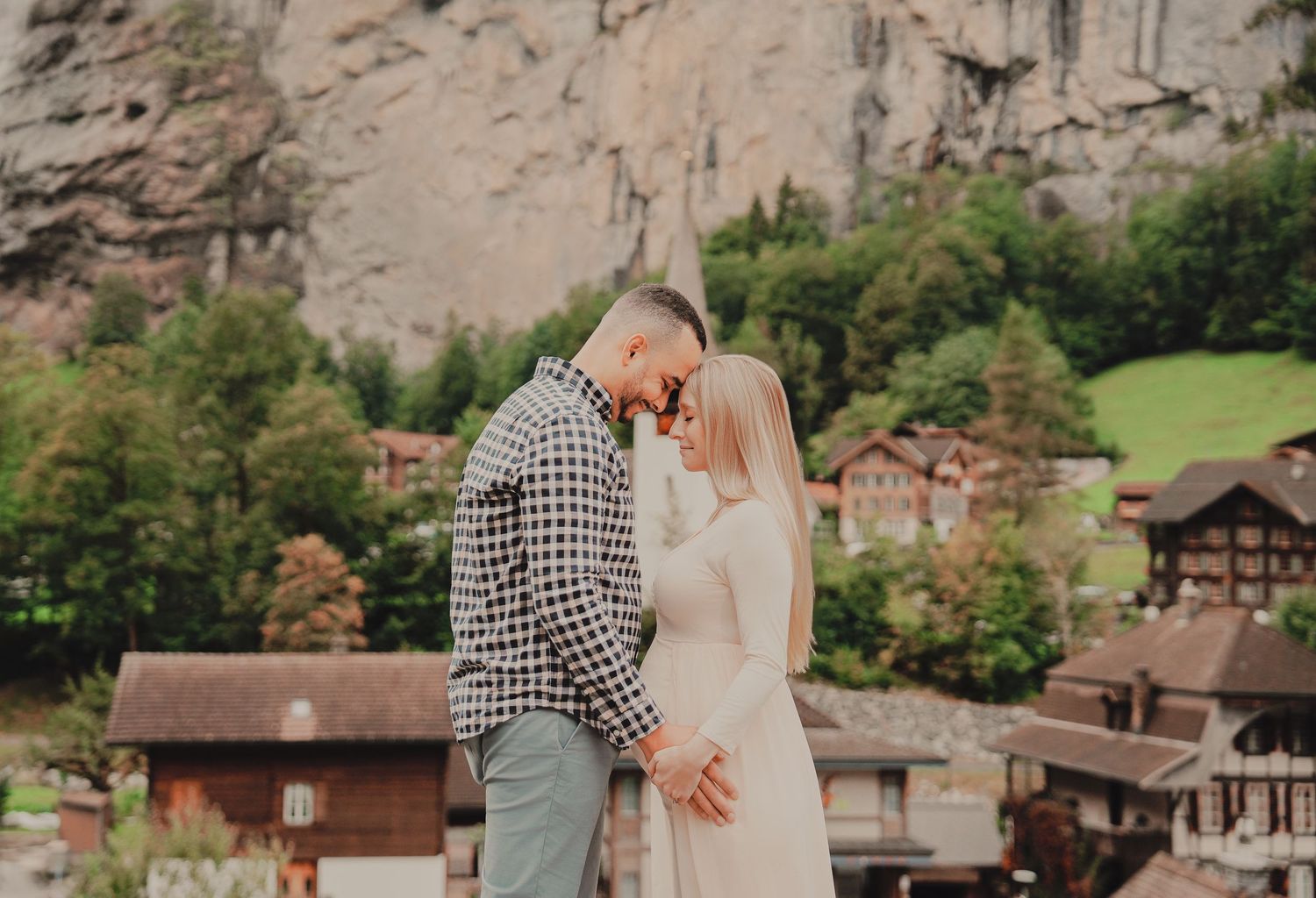 Expecting couple shares intimate forehead touch on bridge with Lauterbrunnen waterfall village below.