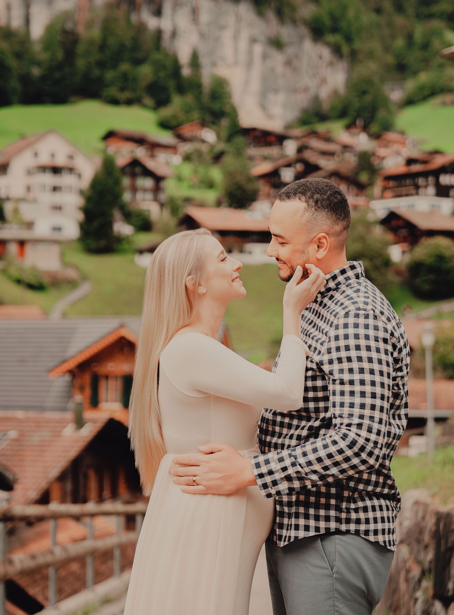 Joyful expecting couple sharing intimate moment with picturesque Swiss alpine village and green hillside behind them.