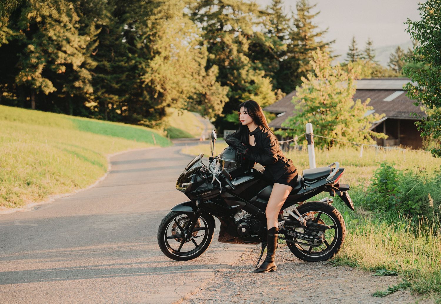 Stylish woman in black mini dress standing next to a black motorcycle on a countryside road surrounded by trees.