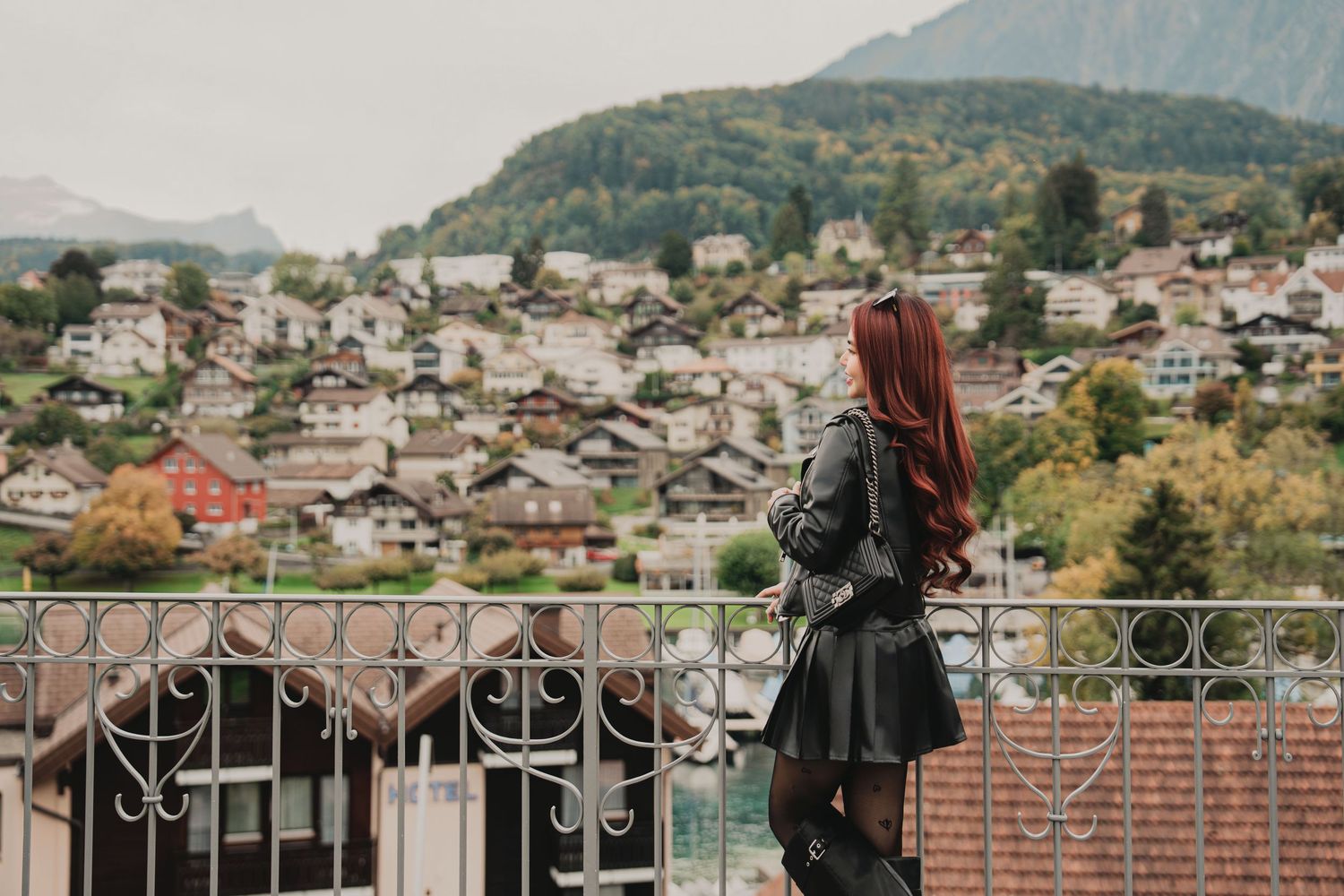 Red-haired woman overlooks a picturesque Swiss village from a decorative iron balcony railing.
