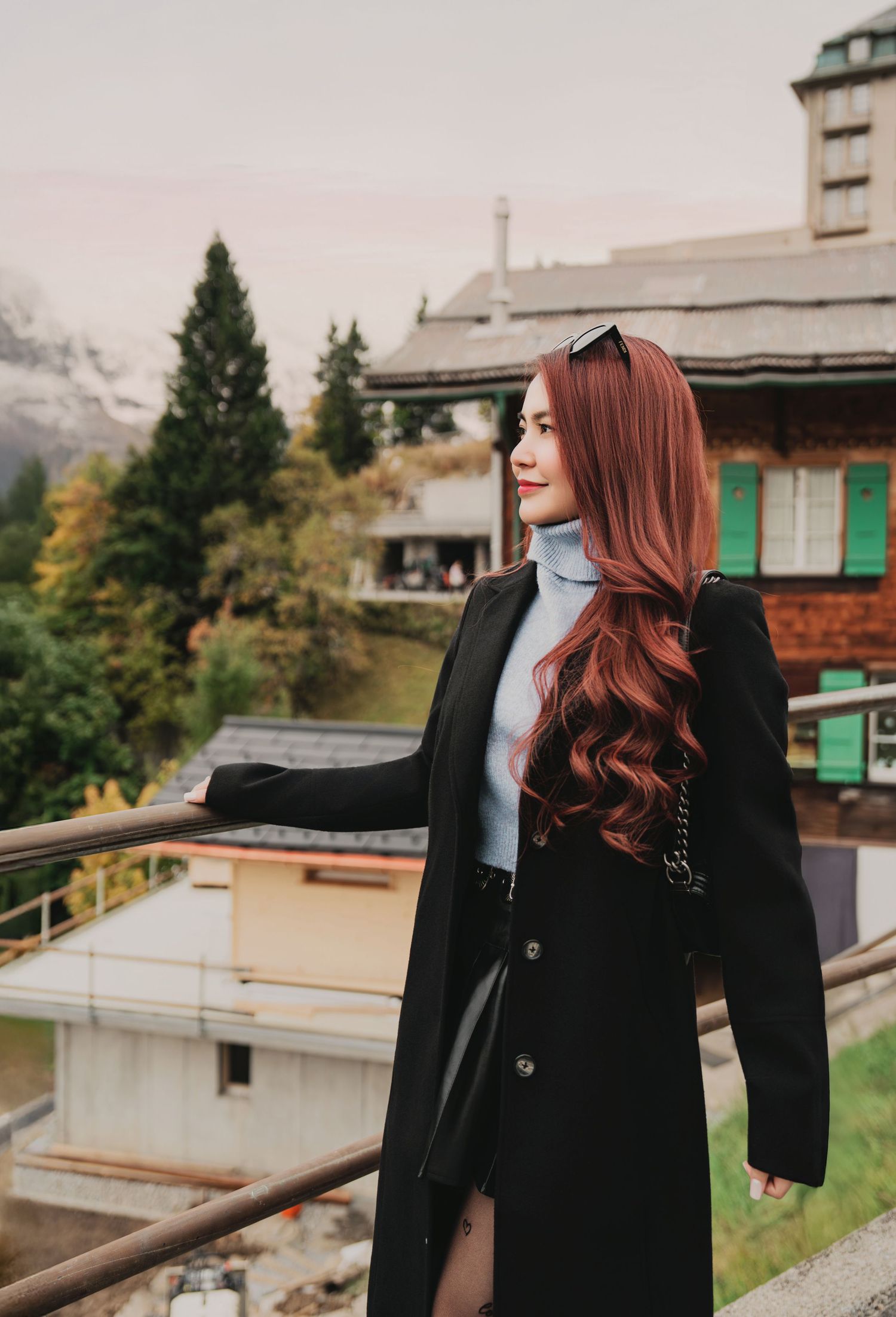 Elegant woman in long black coat and blue turtleneck standing on balcony with Alpine village backdrop.
