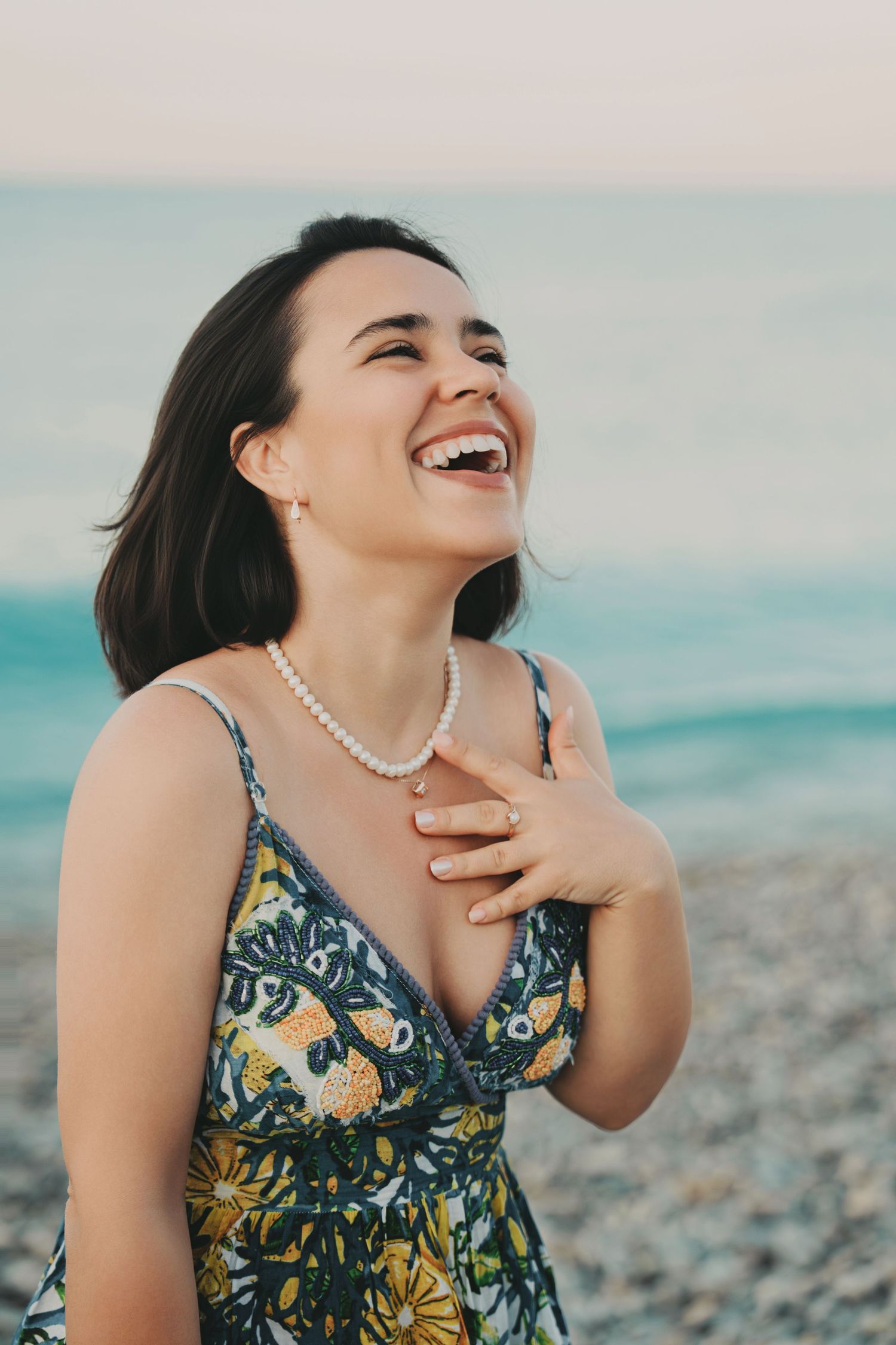 Joyful brunette woman laughing in floral sundress and pearl necklace at beach.
