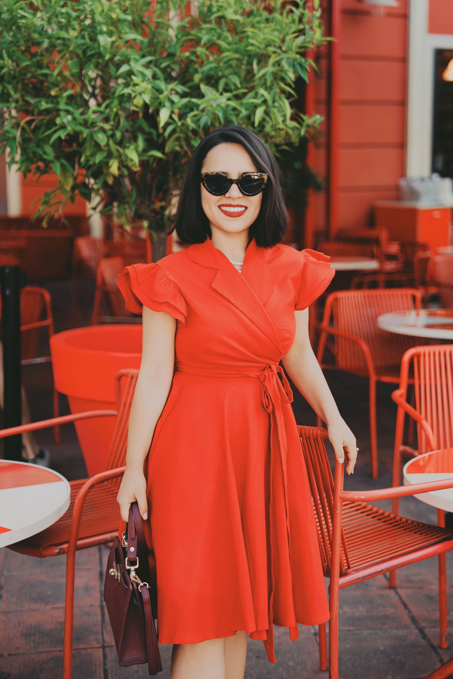 Smiling woman in red wrap dress and sunglasses standing near red outdoor cafe furniture.