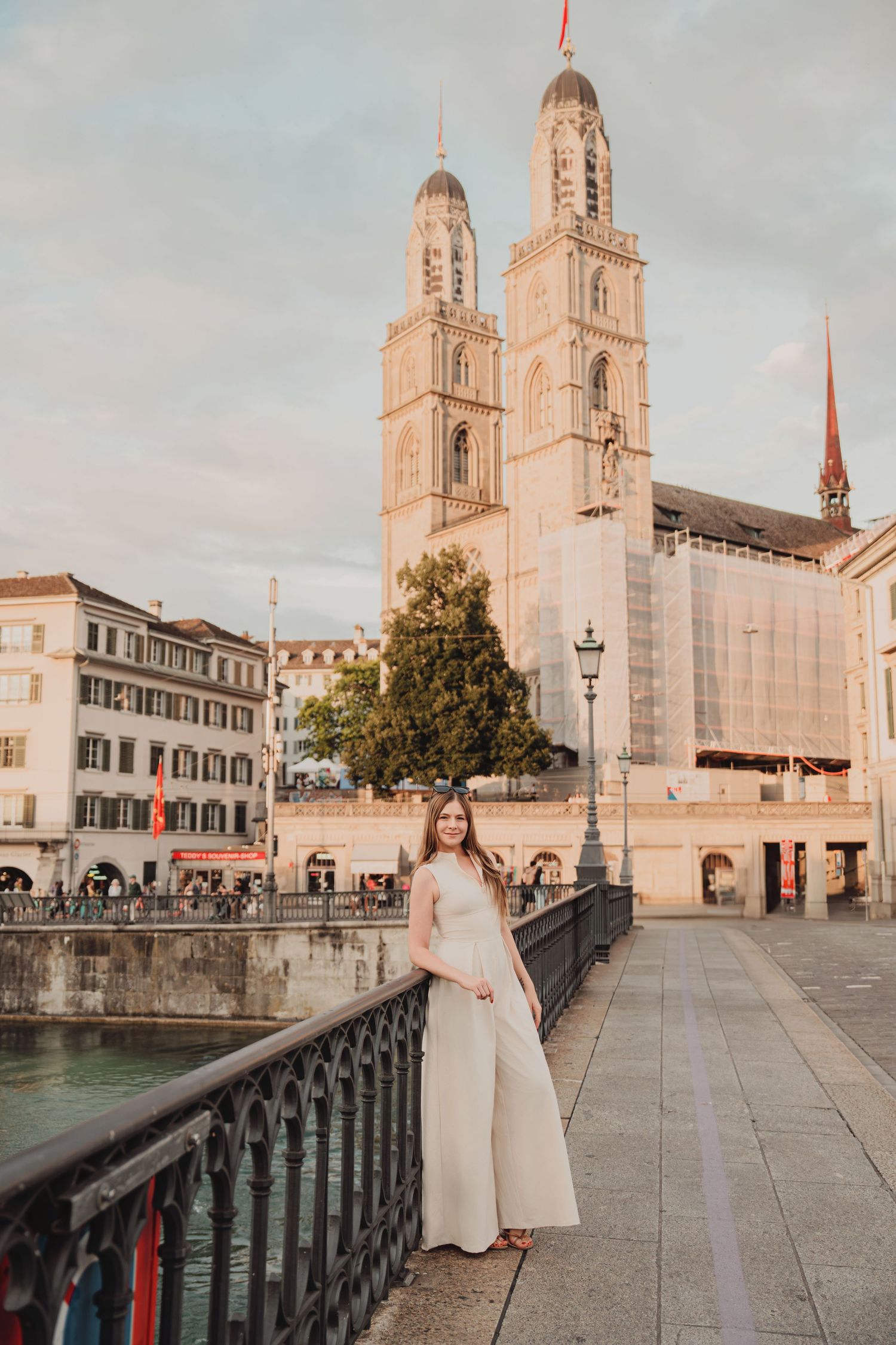 Elegant woman in cream dress stands on bridge with Grossmunster Cathedral twin towers in Zurich.