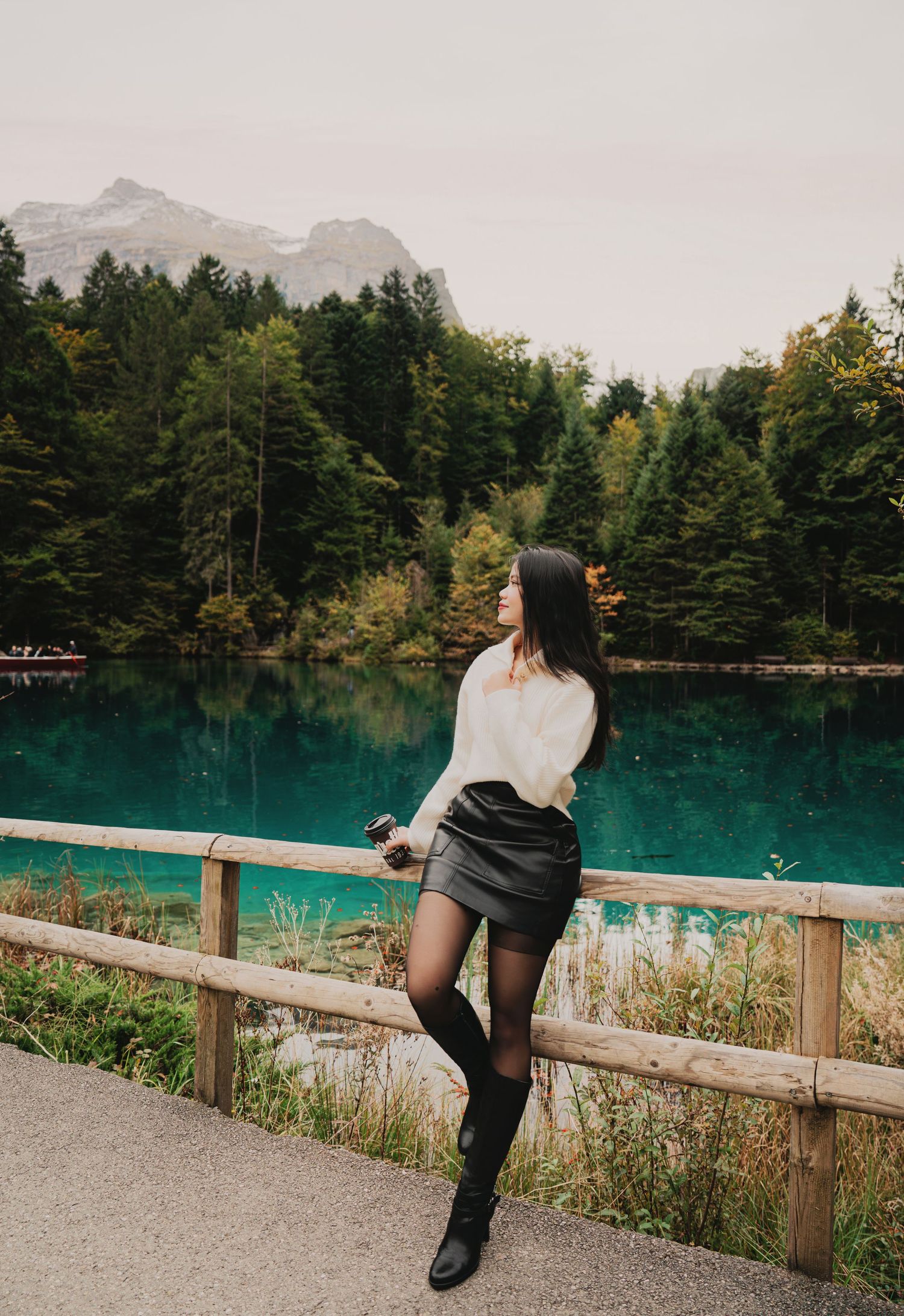 Woman in white sweater and black skirt standing by wooden fence overlooking turquoise alpine mountain lake.