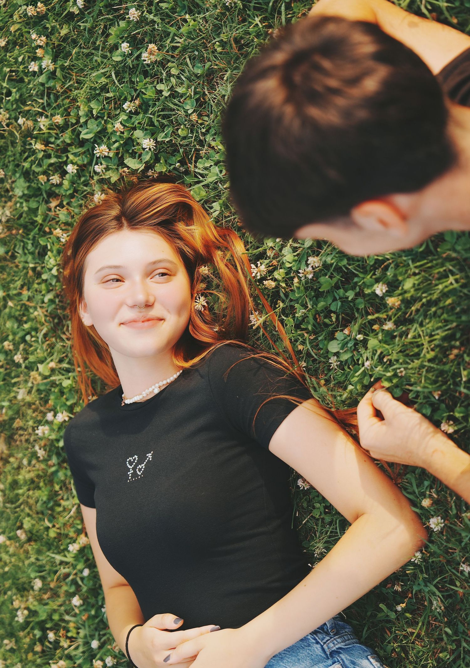 Smiling redhead girl lying on flower-dotted grass while her partner sits beside her.