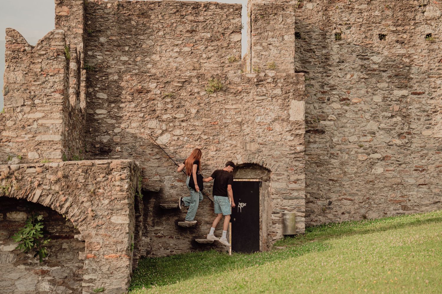 A couple exploring the historic stone walls and entrance of a medieval castle ruin on a summer day.