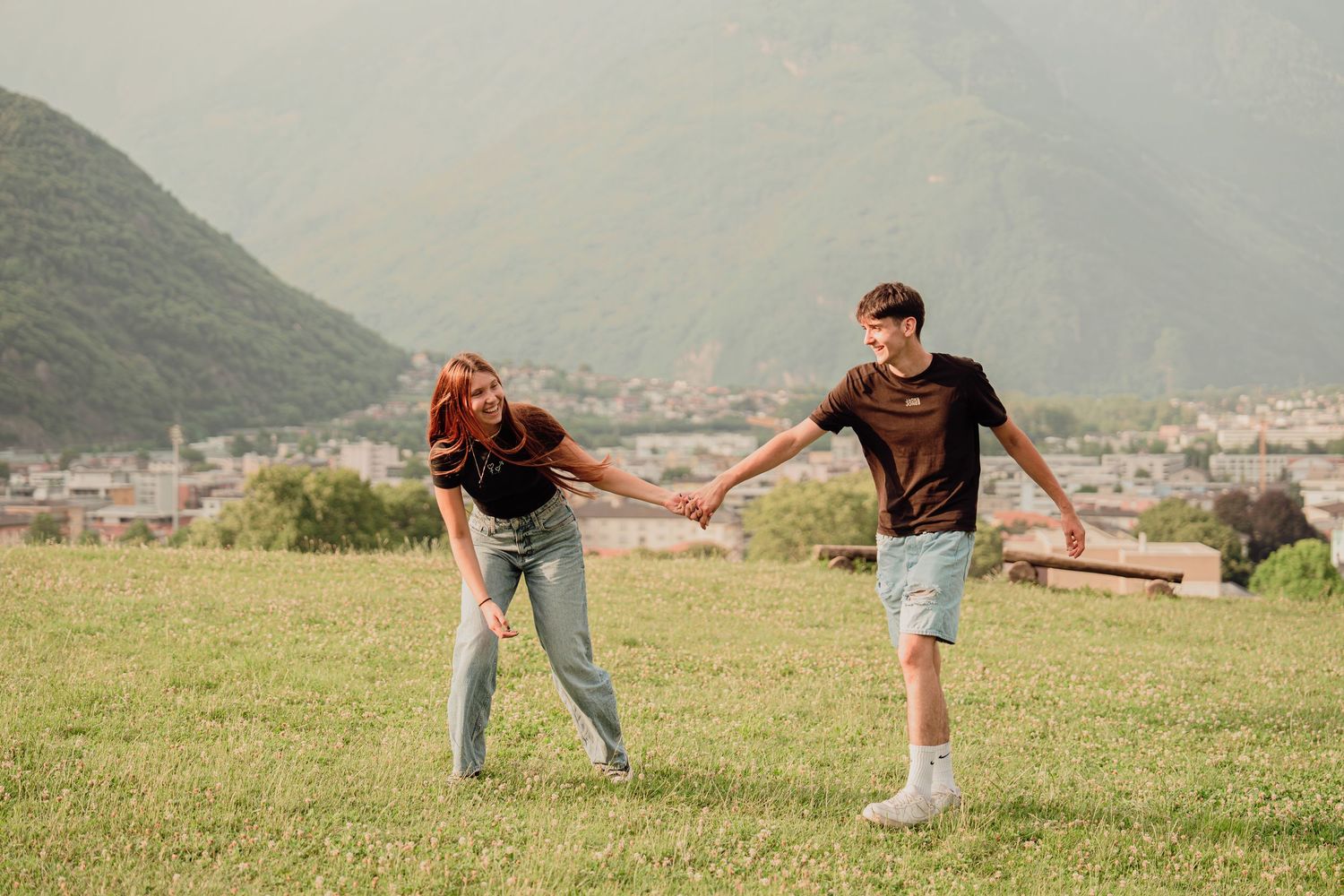 Young couple holding hands on a green meadow with mountain village backdrop.