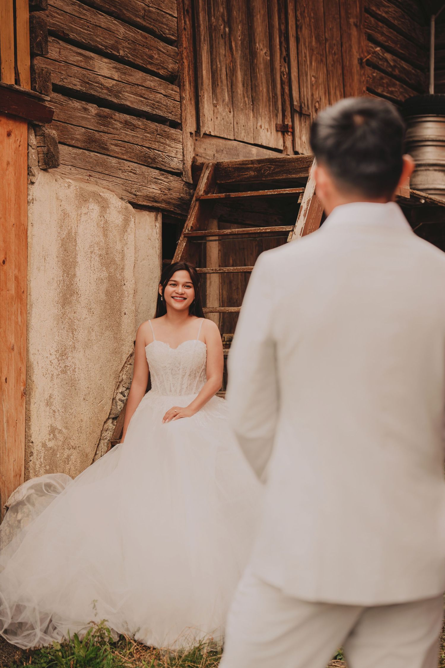 Radiant bride in strapless white ballgown smiles at groom approaching rustic wooden barn steps.