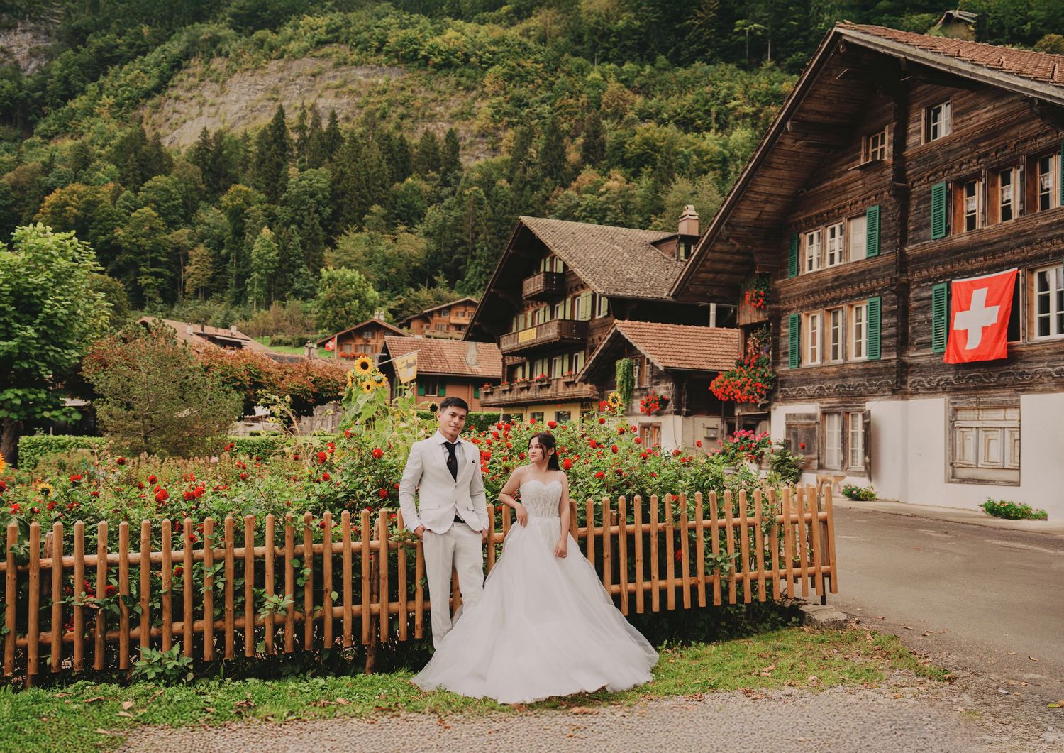 Wedding couple standing before a traditional Swiss chalet with red flowers, wooden fence, and Swiss flag.
