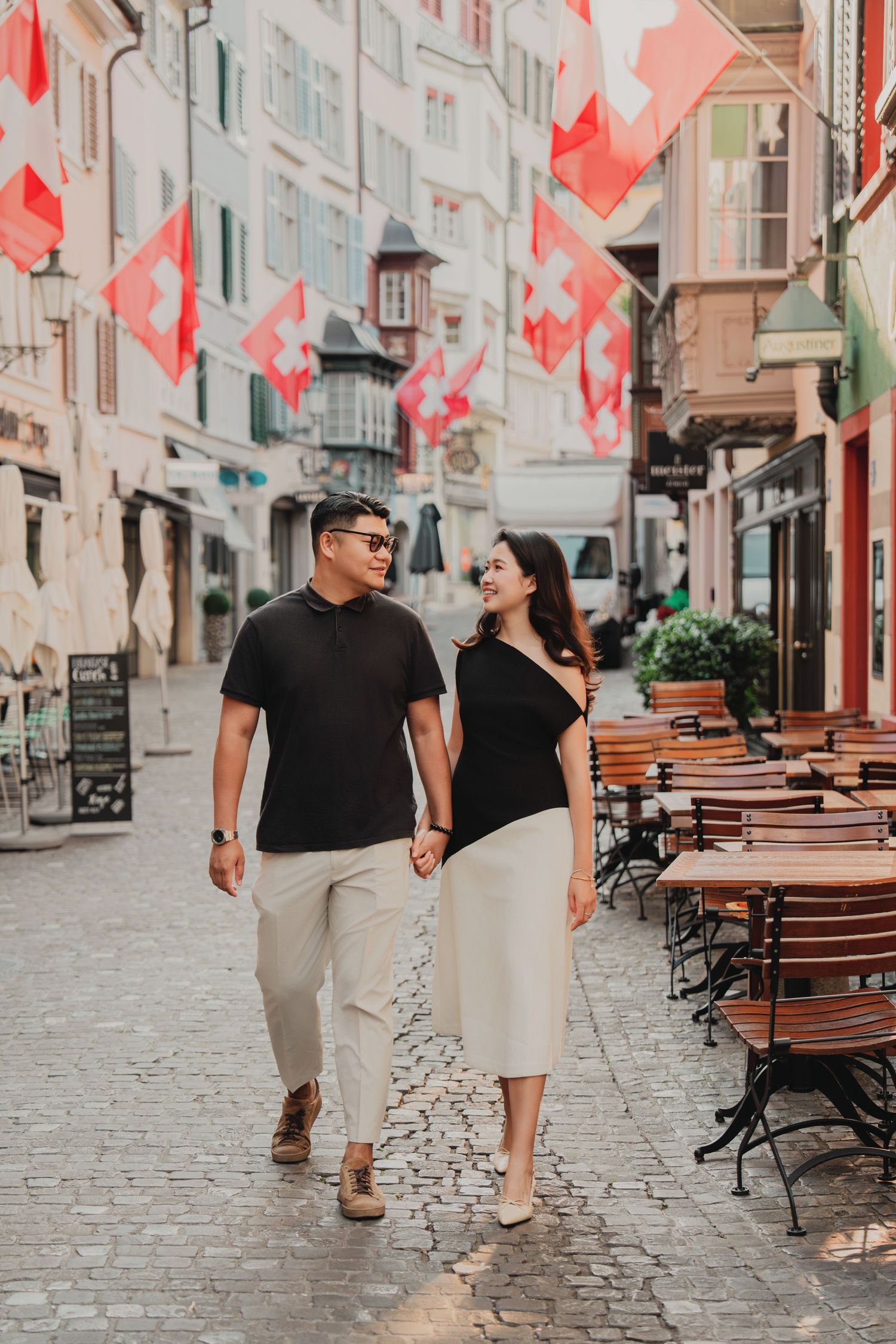 Smiling couple walking together on a cobblestone street in Zurich with Swiss flags hanging above.