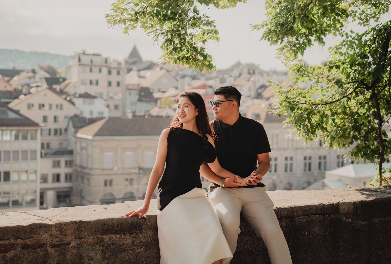 Couple sitting together on a stone wall with a scenic Zurich cityscape and green trees behind them.