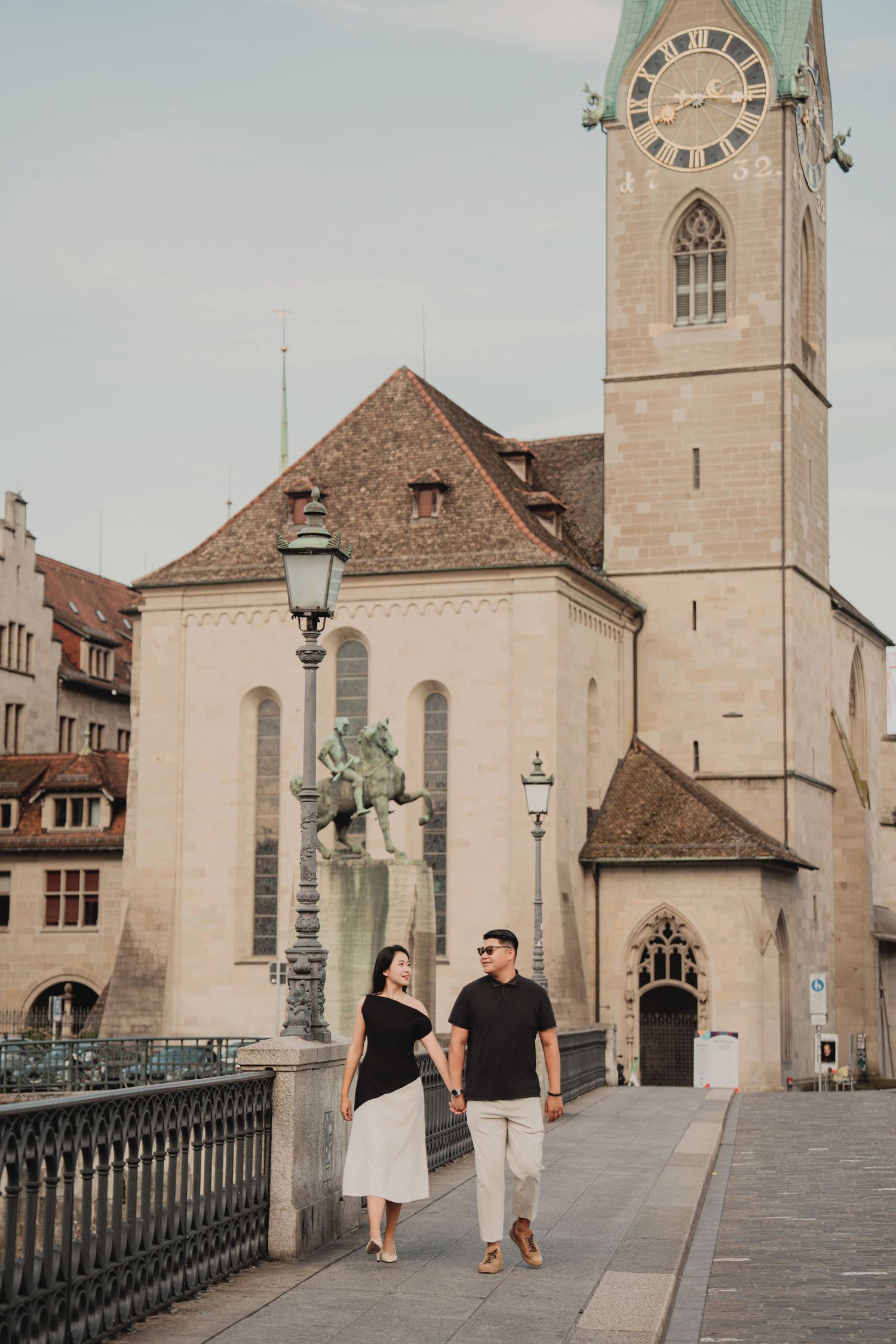 Couple holding hands walking on a bridge in Zurich's old town with Fraumunster Church tower behind.