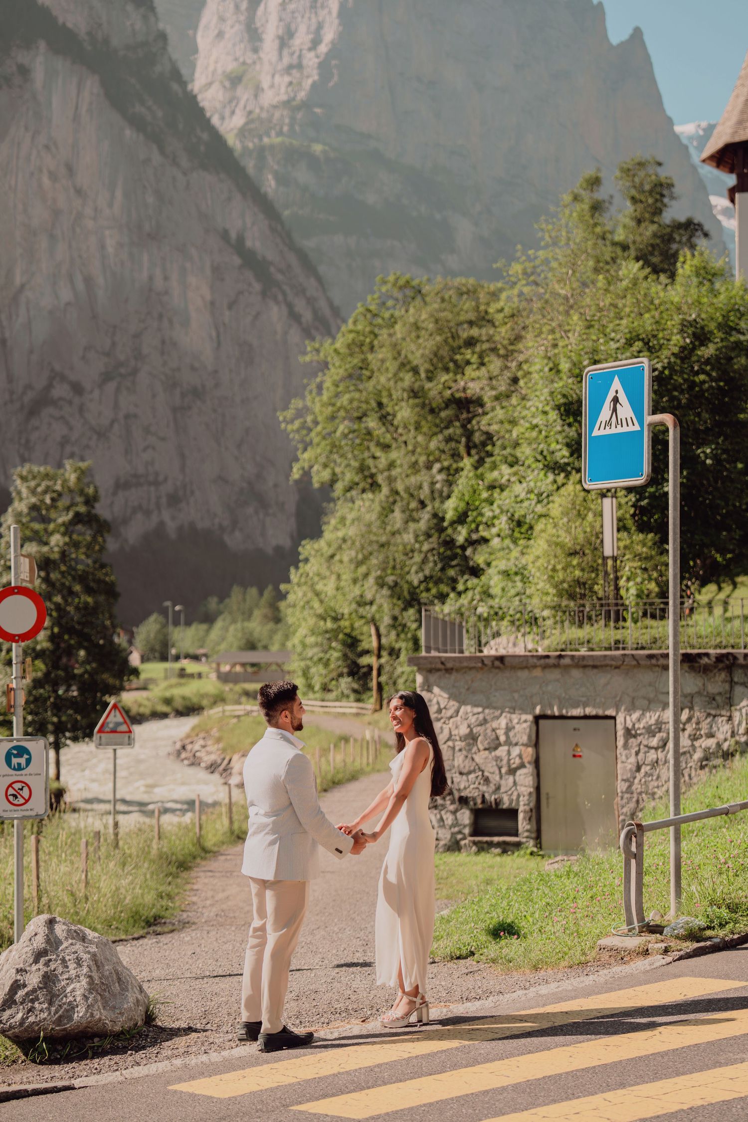 Couple holding hands at a crosswalk in Lauterbrunnen, Switzerland, with dramatic mountain cliffs in the background.