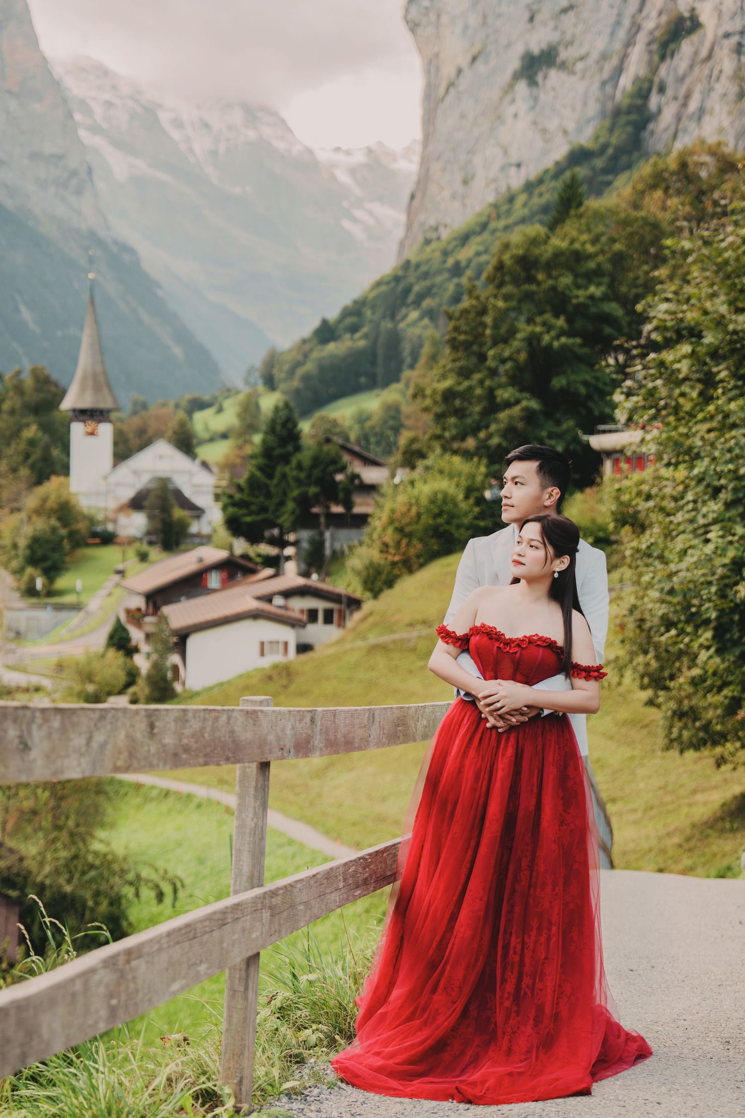 Couple embracing on a bridge in red gown and grey suit with Lauterbrunnen village and Alps in the background.