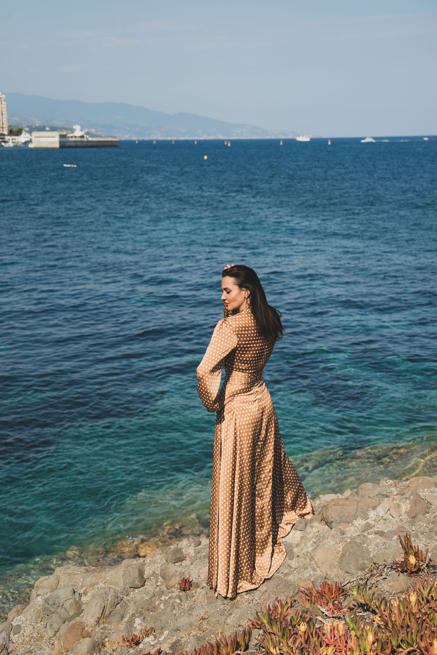 Elegant woman in polka dot dress stands on rocky shore gazing at blue ocean horizon.