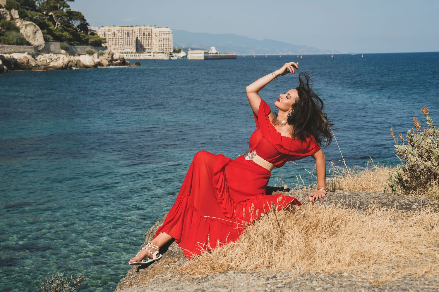 Brunette woman in red two-piece outfit posing on rocky hillside with historic coastal fortress backdrop.