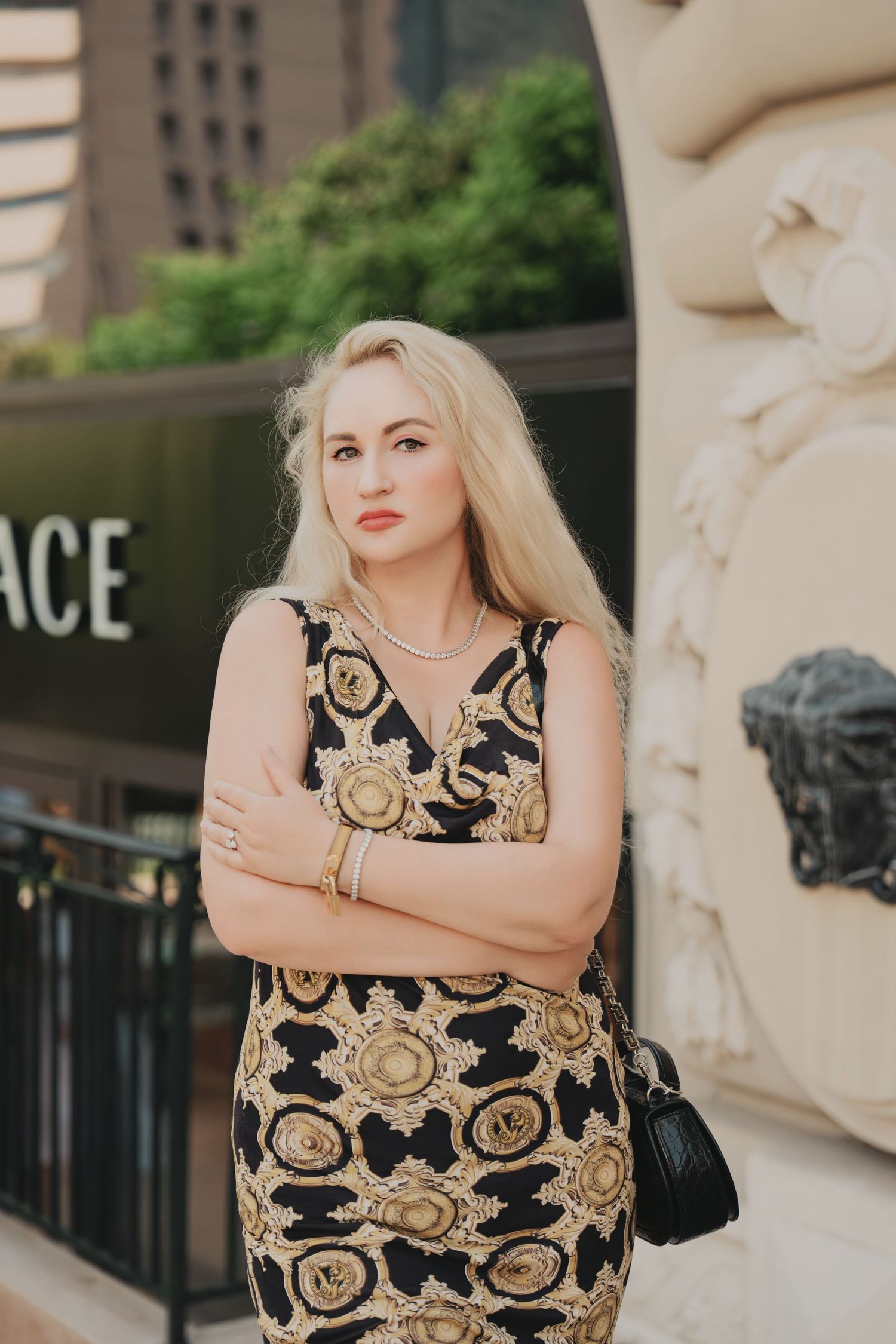 Elegant blonde woman in ornate black and gold Versace dress standing outside luxury boutique.