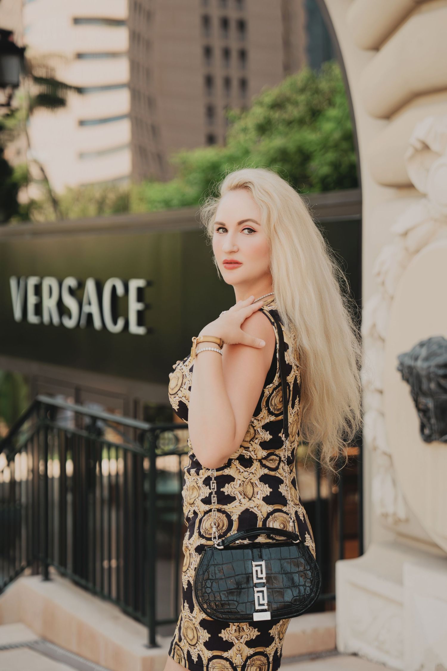 Blonde woman in Versace print dress holding designer bag outside luxury Versace boutique.