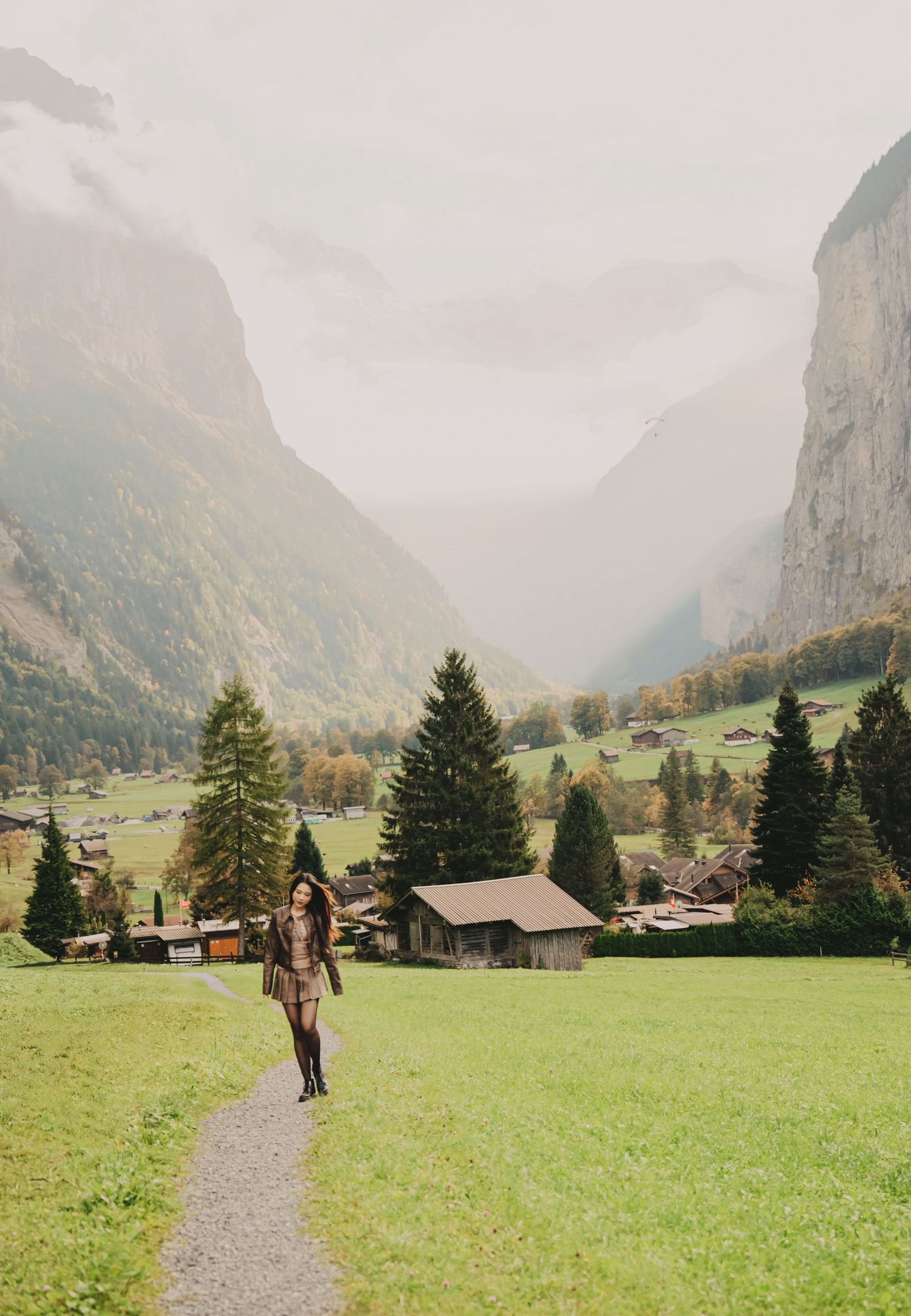 Woman walking on a gravel path through a lush green Swiss valley with towering misty mountains.