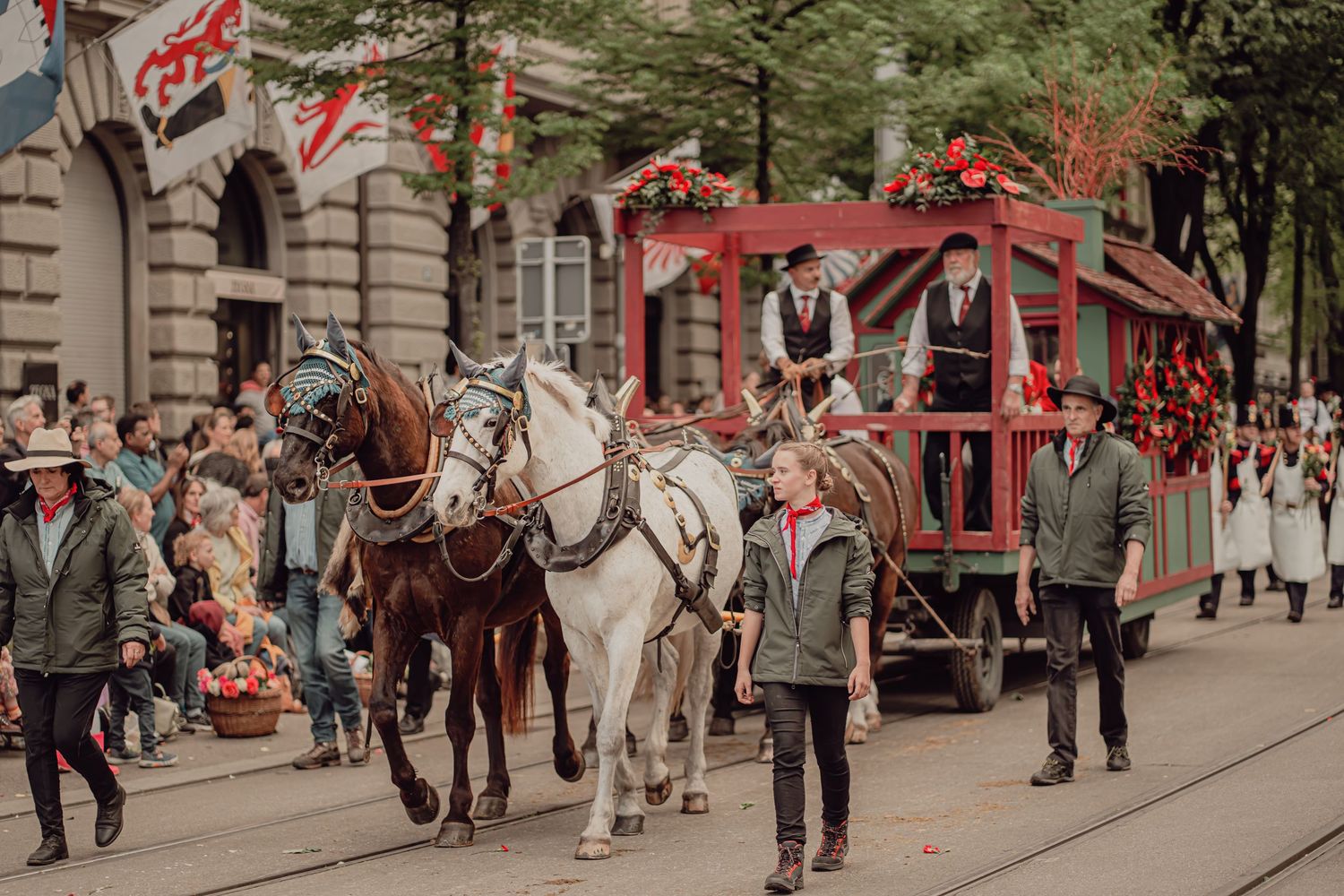 A decorated red horse-drawn carriage leads a traditional Swiss parade through city streets.