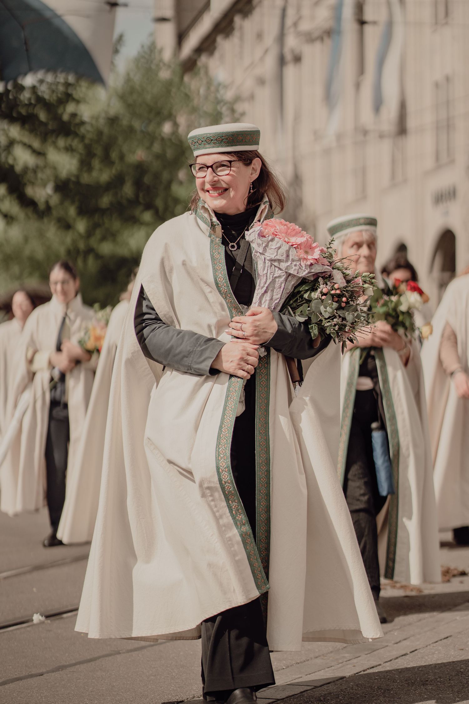 Smiling woman in white ceremonial robe and green hat holding a bouquet of flowers during a parade.