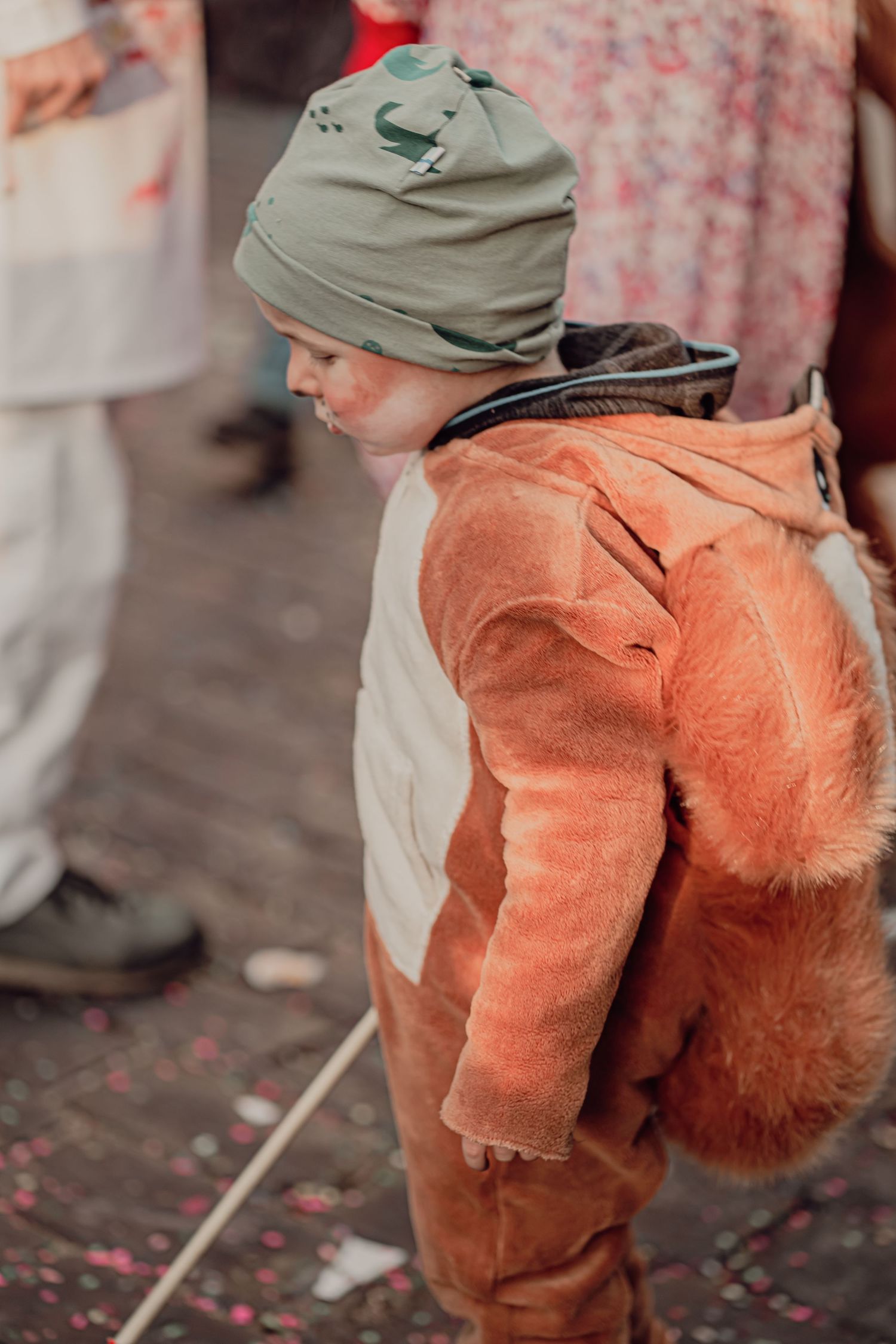 Toddler in orange fox costume with green hat at colorful carnival parade.