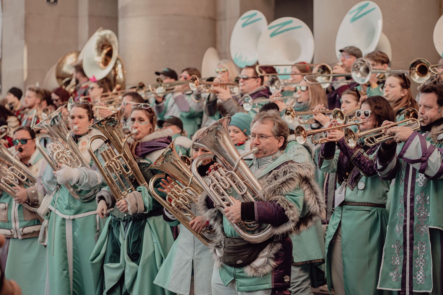 A marching band in teal and purple medieval costumes plays brass instruments at a parade.