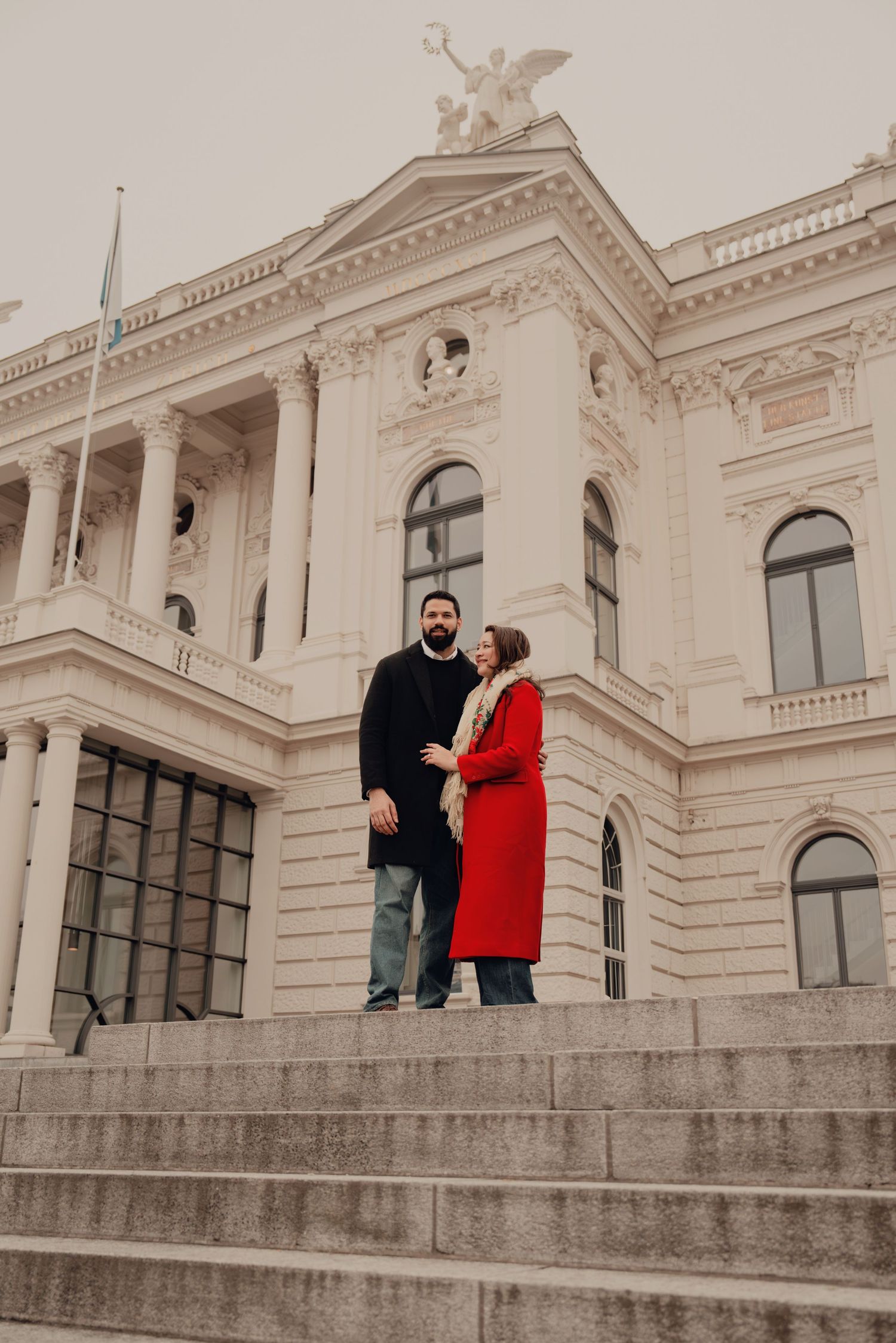 Couple stands close on steps of a grand white neoclassical building on an overcast day.