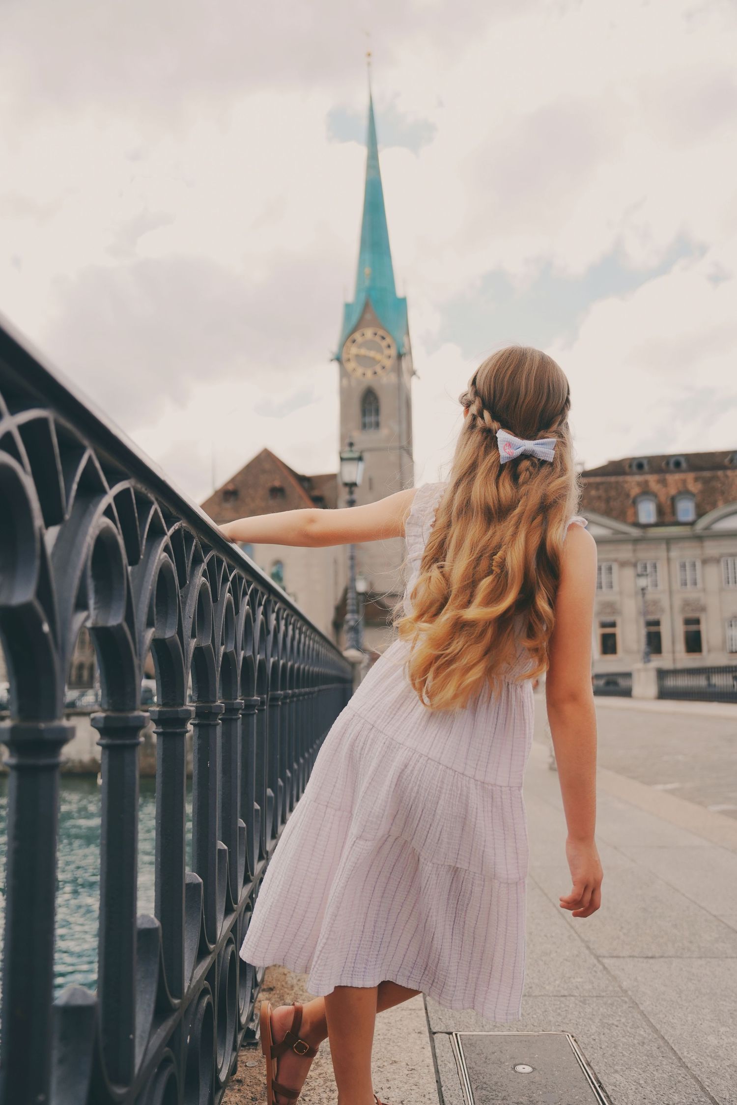 A young blonde girl in a white dress walks along a black iron bridge railing toward Fraumunster church in Zurich.