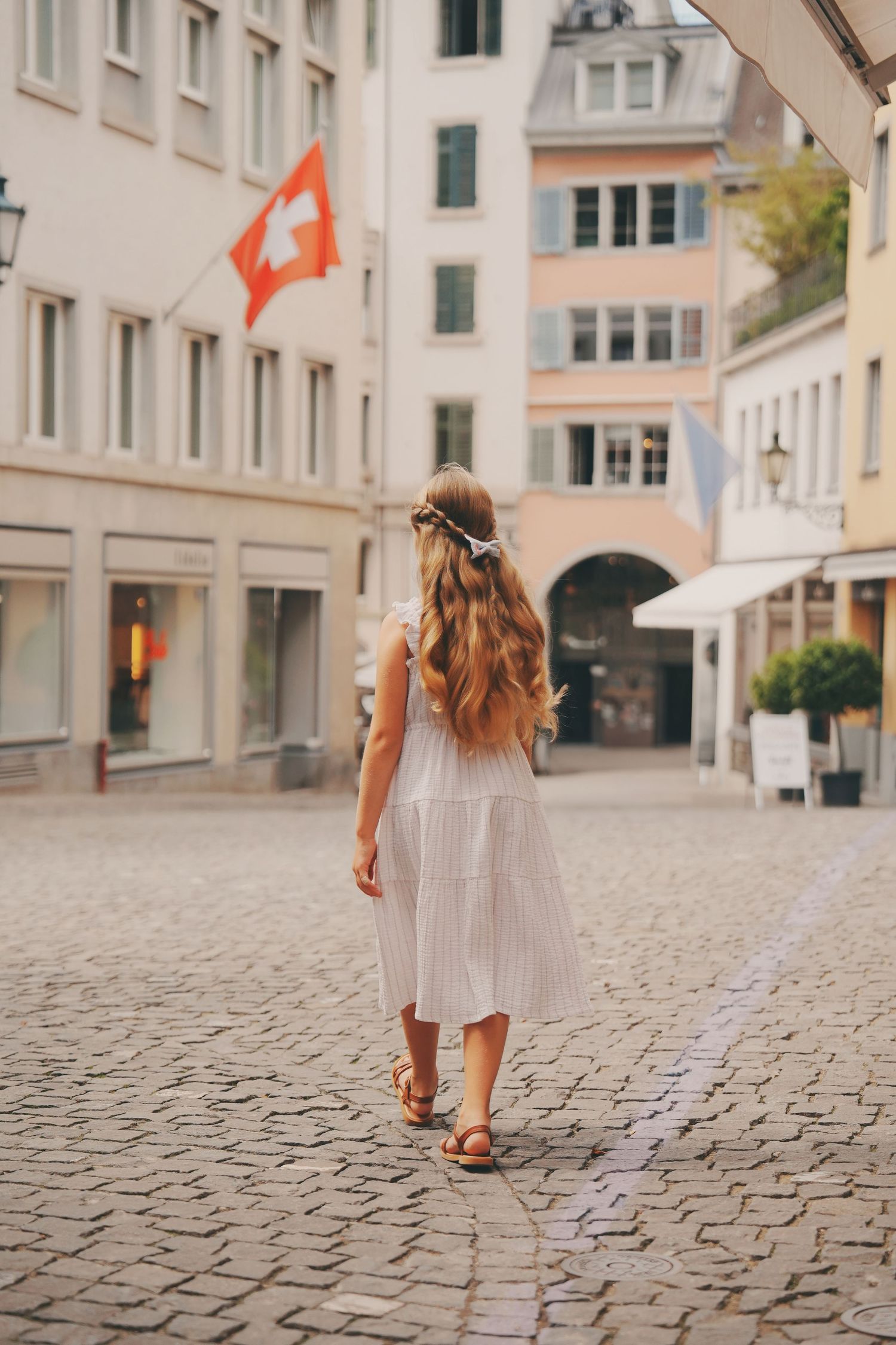 Young girl in white dress with flower crown walking cobblestone street with Swiss flag backdrop.