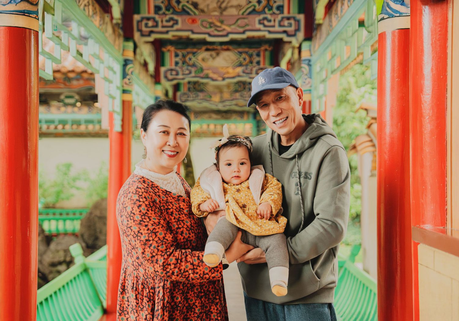 Grandparents smiling with a baby in yellow inside a colorful Chinese temple corridor.