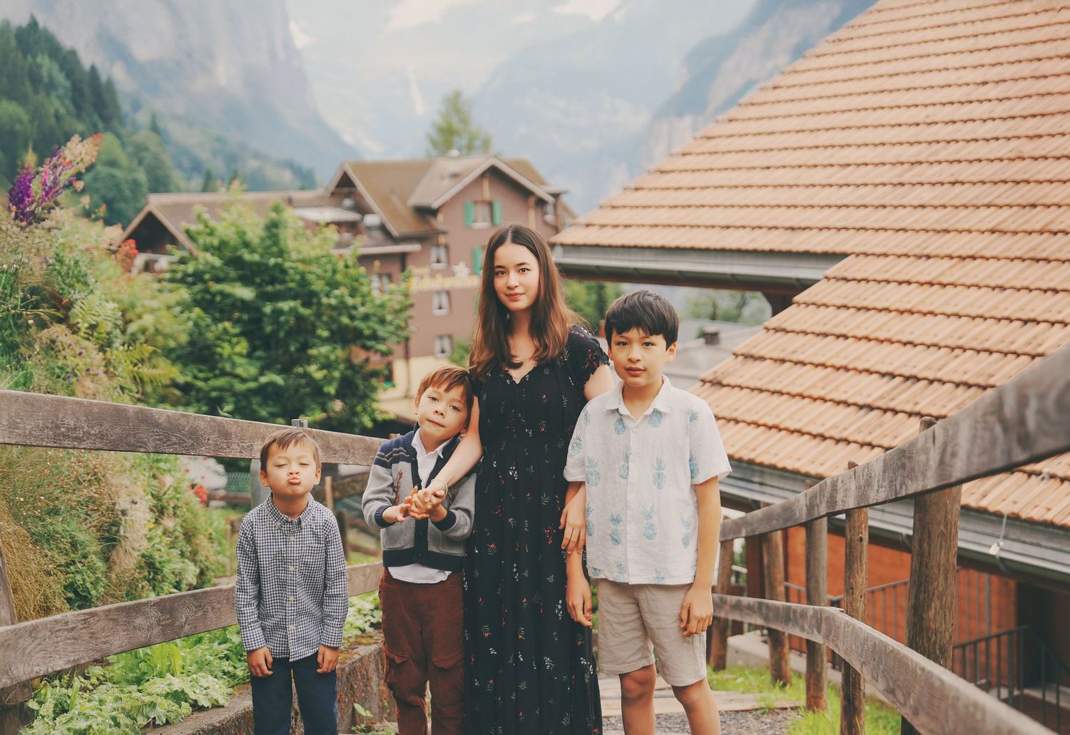 Asian mother stands with three sons on wooden bridge with charming Swiss alpine village backdrop.