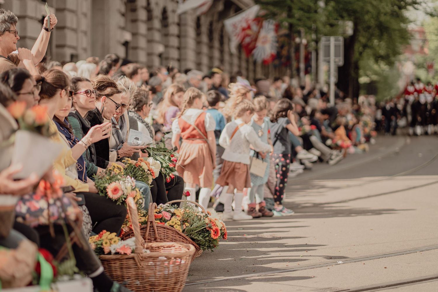 Crowds line a sunny street watching a traditional parade, with flower baskets in the foreground.