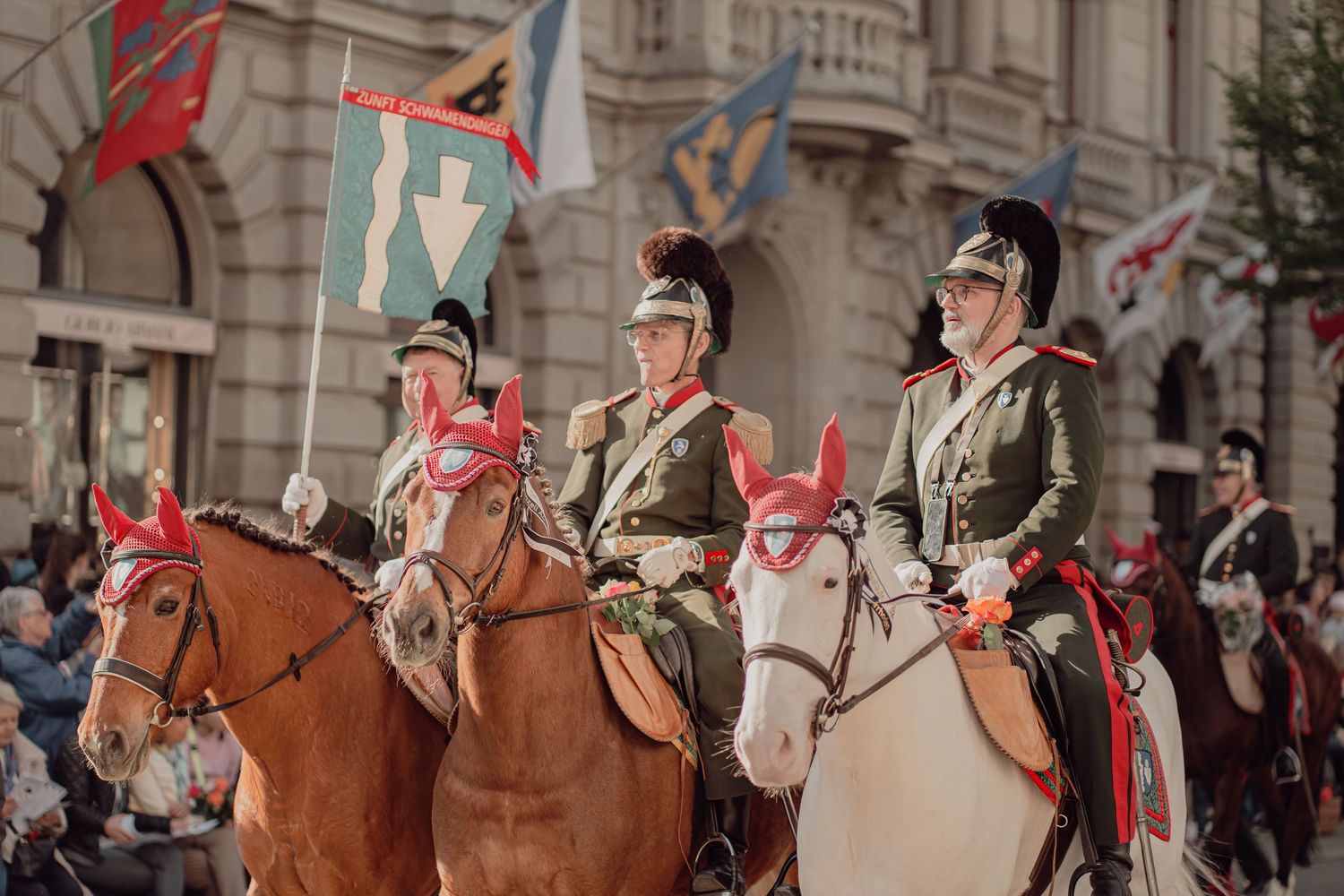 Soldiers in historical uniforms ride horses during a traditional parade with colorful flags waving in the background.