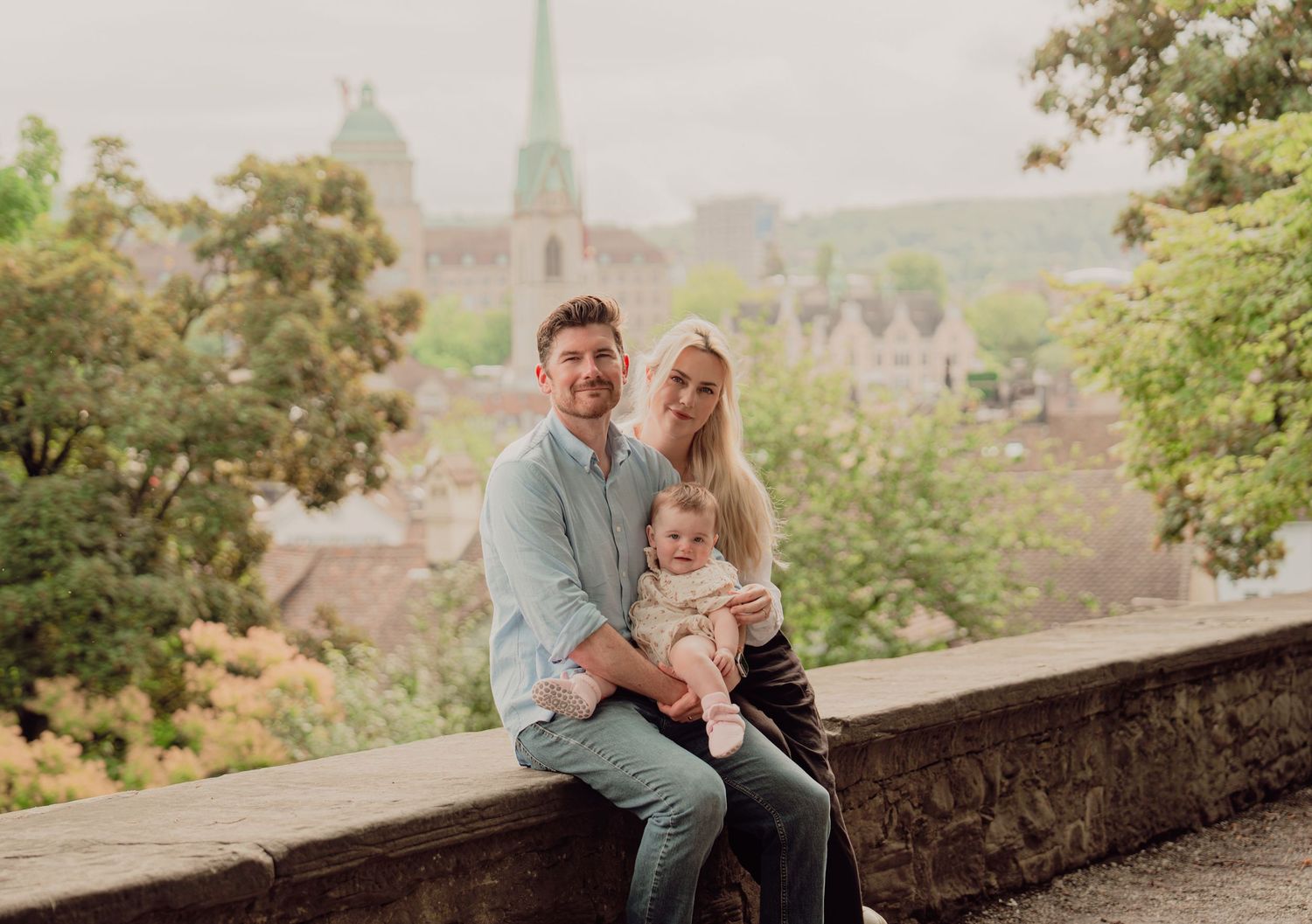 Smiling couple holding baby seated on stone wall with European old town skyline in background.