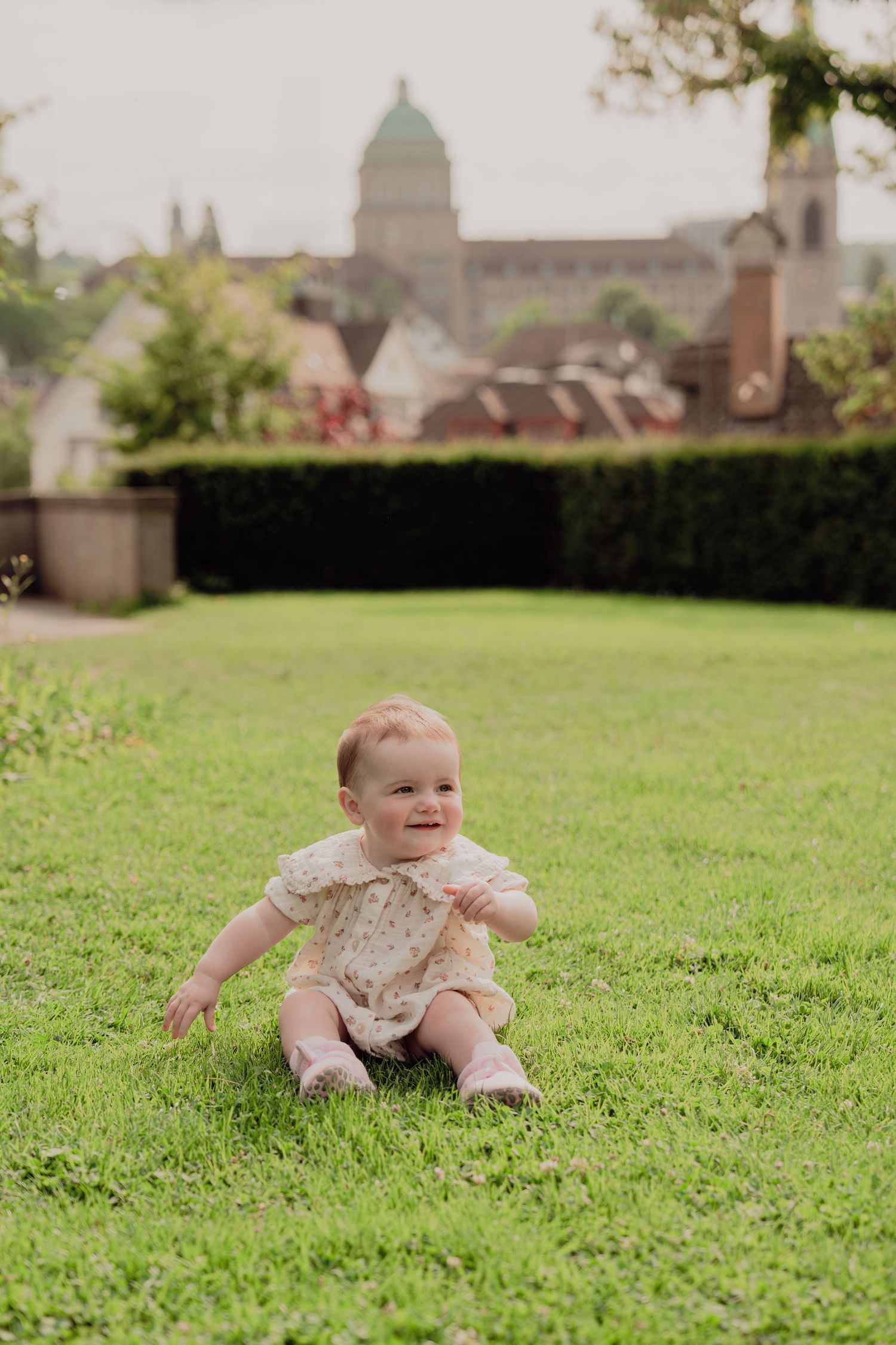 Smiling baby in floral romper sitting on lush green grass with European cityscape behind.