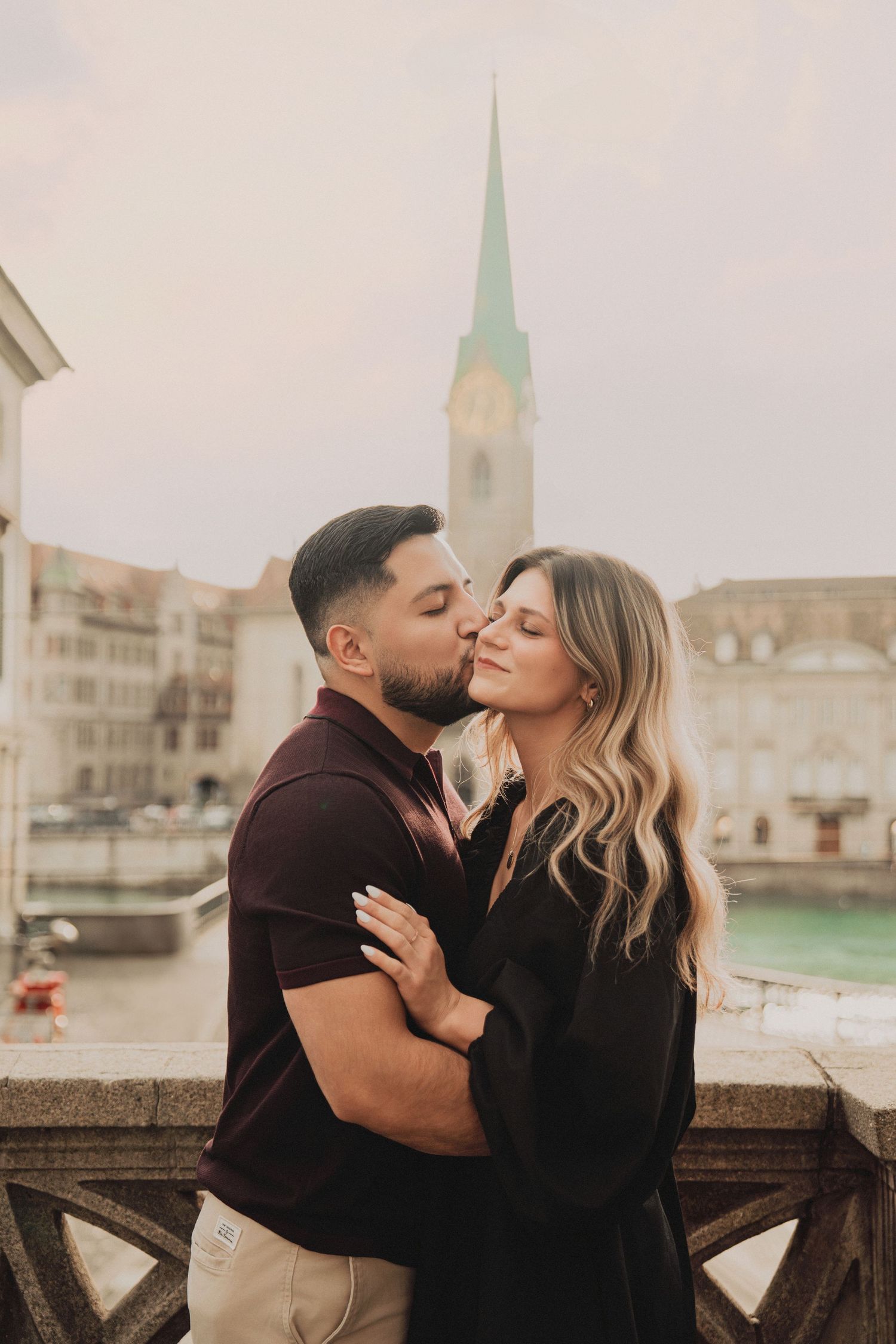 Man kissing woman on cheek on Zurich bridge with Fraumunster spire and river in background.