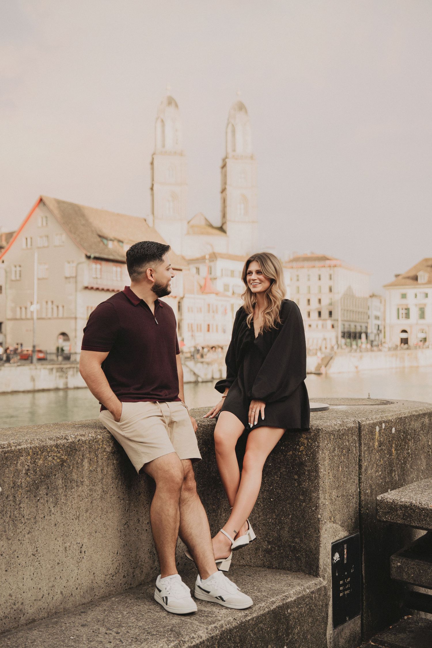 Couple sitting on a stone wall by the Limmat River with Grossmünster church in the background, Zurich.