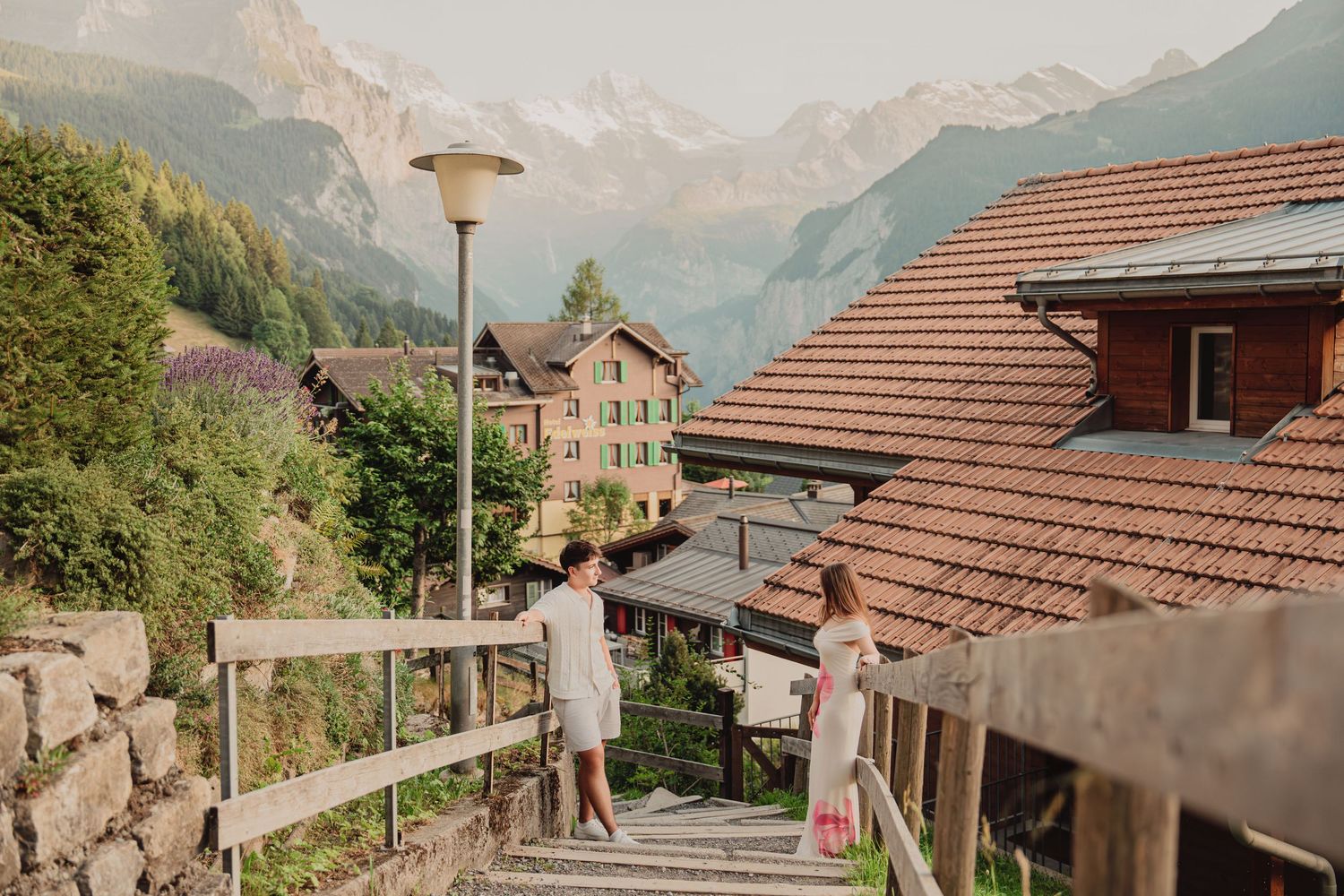 Couple on wooden stairs in a charming Swiss mountain village with colorful buildings and Alpine peaks in background.