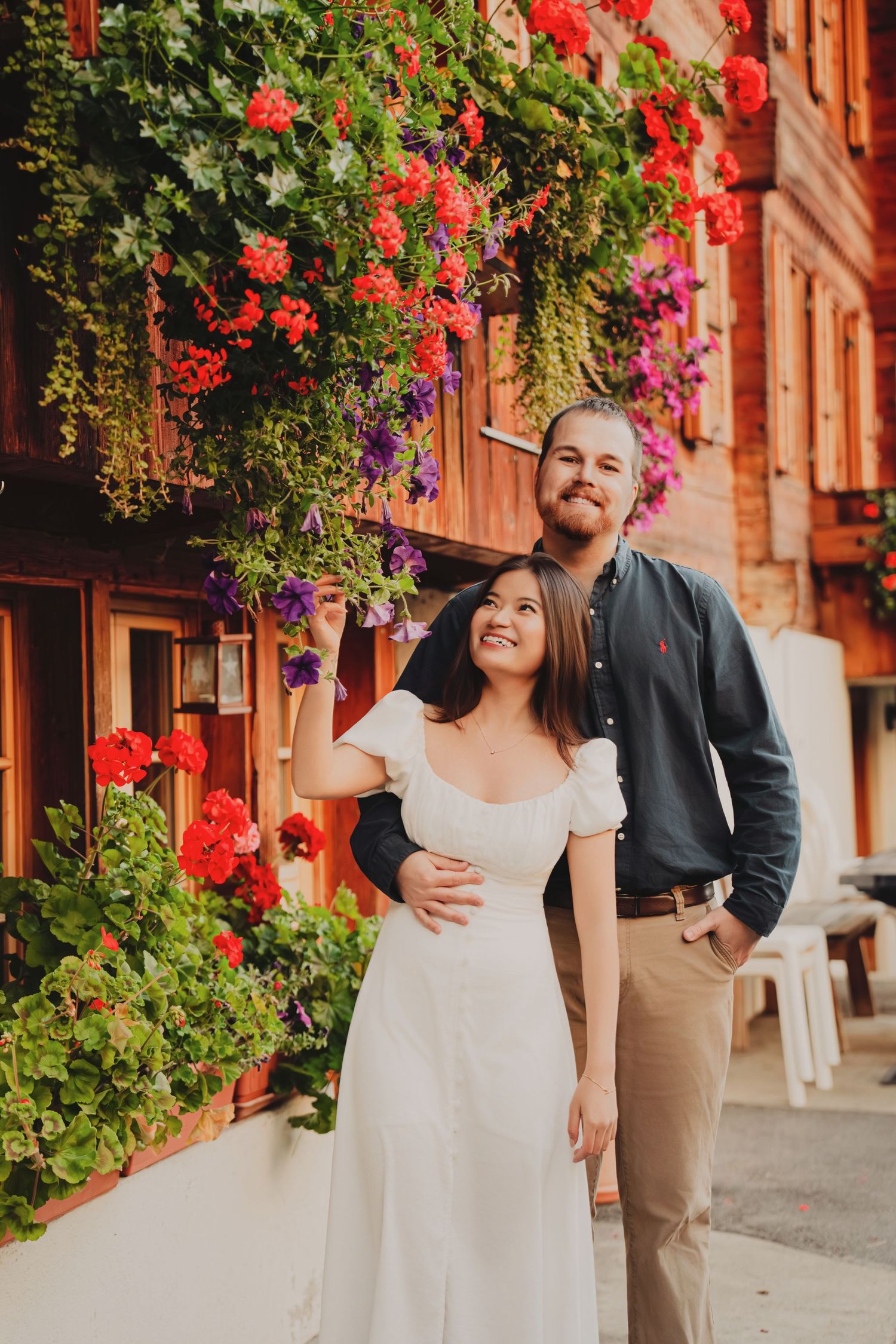 Happy couple standing together under lush red and purple flowers on a Swiss building.