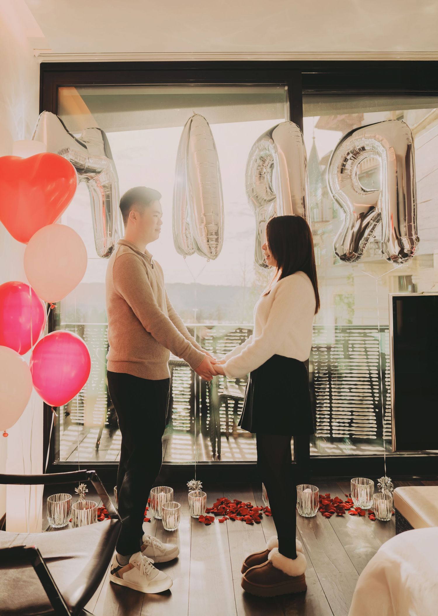 Couple holding hands in romantic room decorated with heart balloons and silver number balloons for proposal.