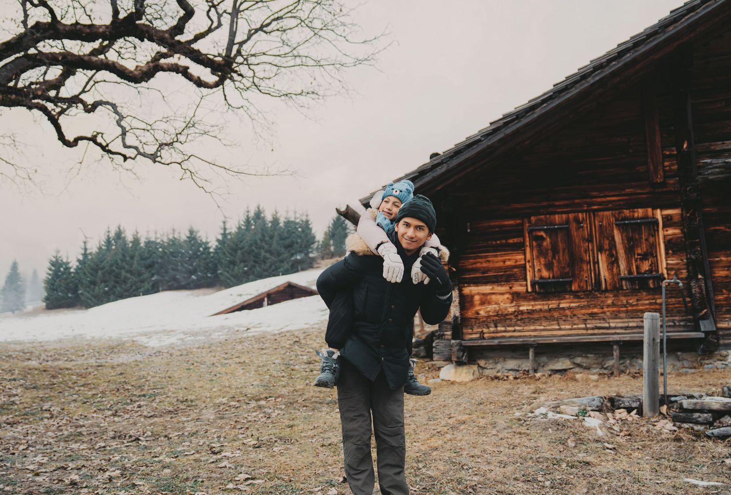 Parent lifting smiling child in blue hat outside rustic wooden cabin in snowy alpine setting.